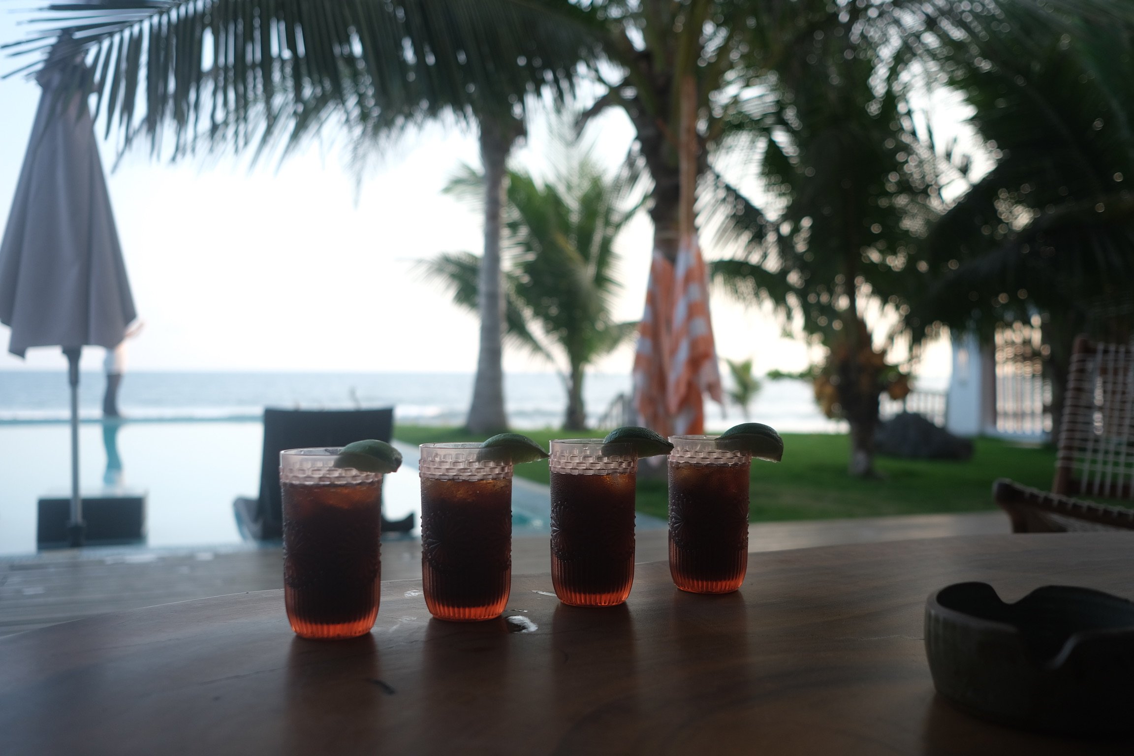 Four glasses of dark-colored drinks with lime wedges on a wooden table, set against a backdrop of palm trees, a swimming pool, and the ocean at sunset or dusk.