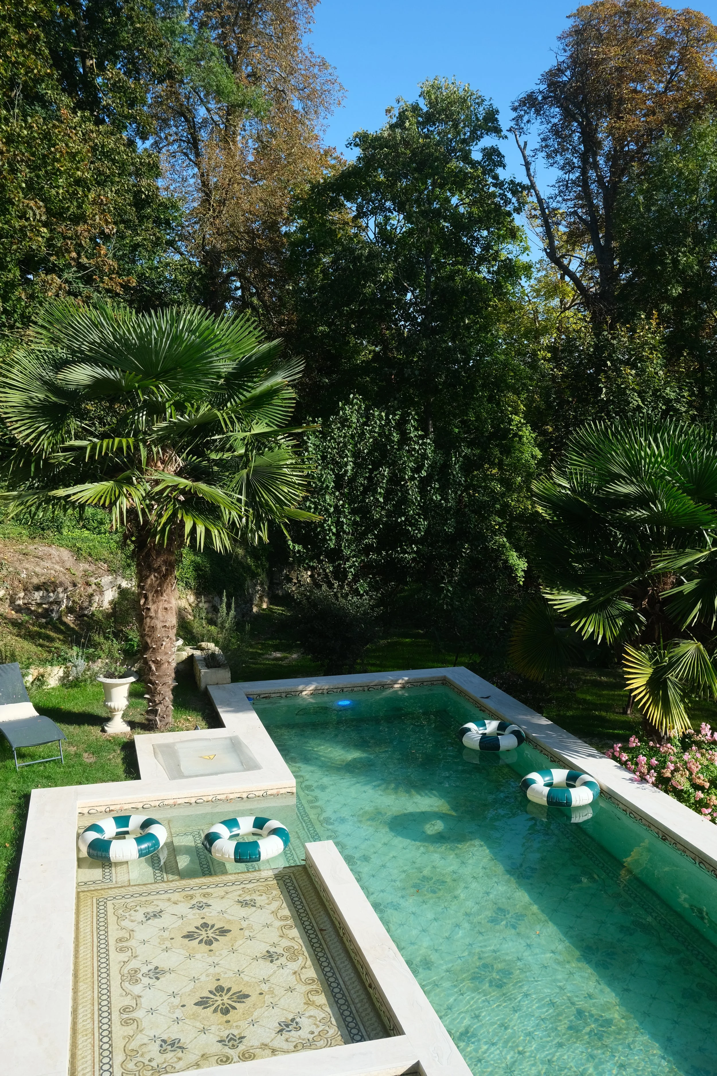 A backyard with a rectangular swimming pool surrounded by lush green trees, tropical plants, and pink flowers. There's a lounge chair, a white planter, and a tiled area near the pool.