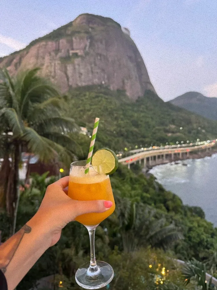 A hand holding a cocktail with lime wedge and straw, overlooking Sugarloaf Mountain and a coastline at dusk.