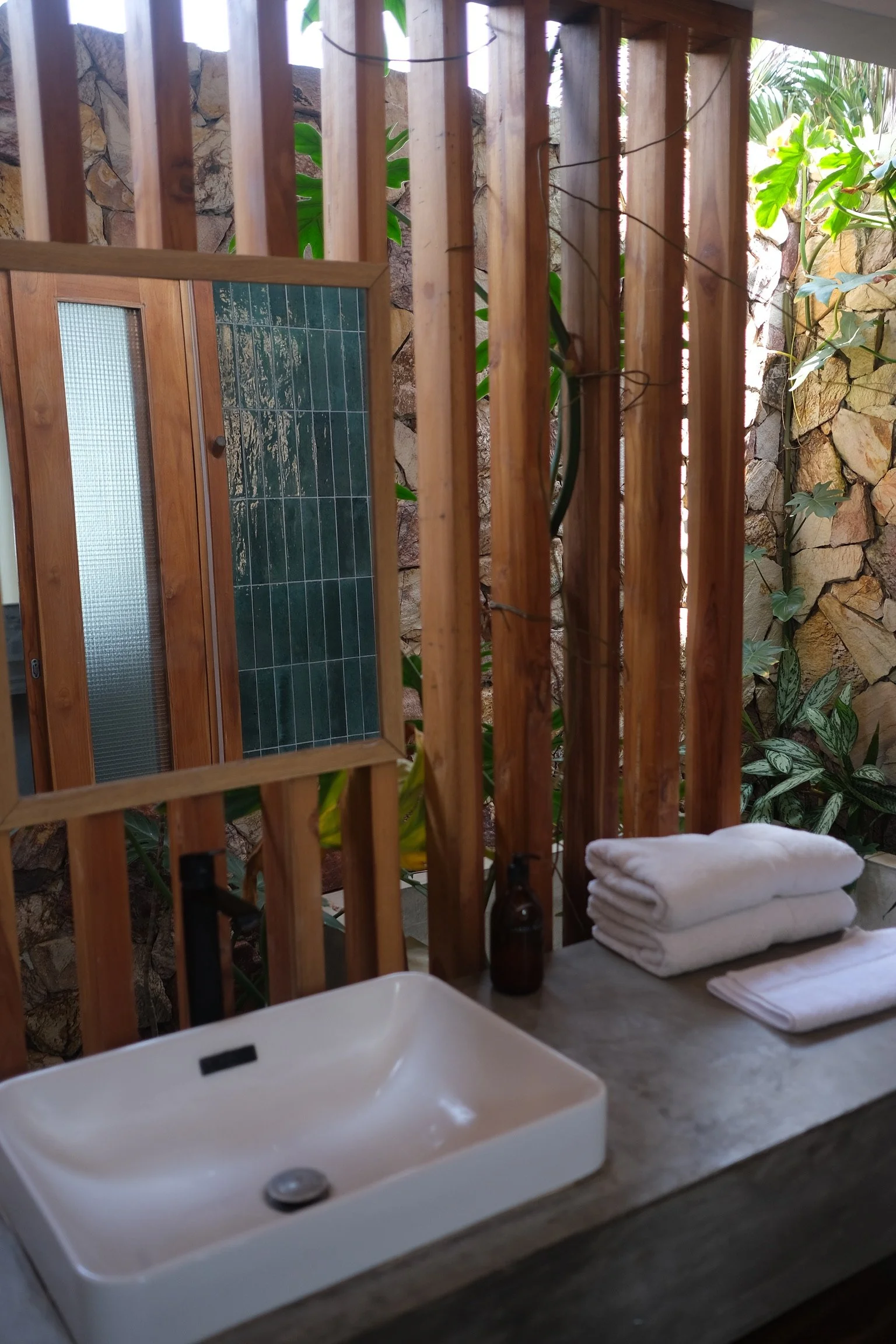 A bathroom countertop with a white ceramic sink, folded white towels, small bottles, and a mirror reflected in a wooden frame, set against a backdrop of green plants and a stone wall.