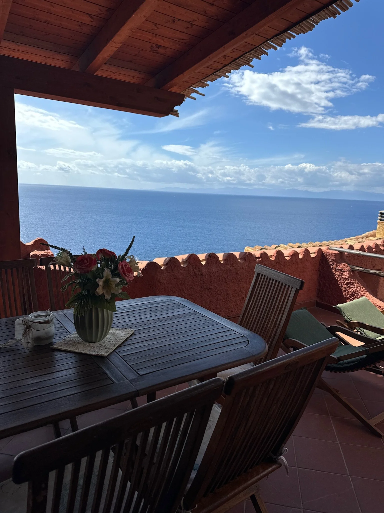 A balcony with a wooden table and chairs, overlooking the ocean with a cloudy sky.