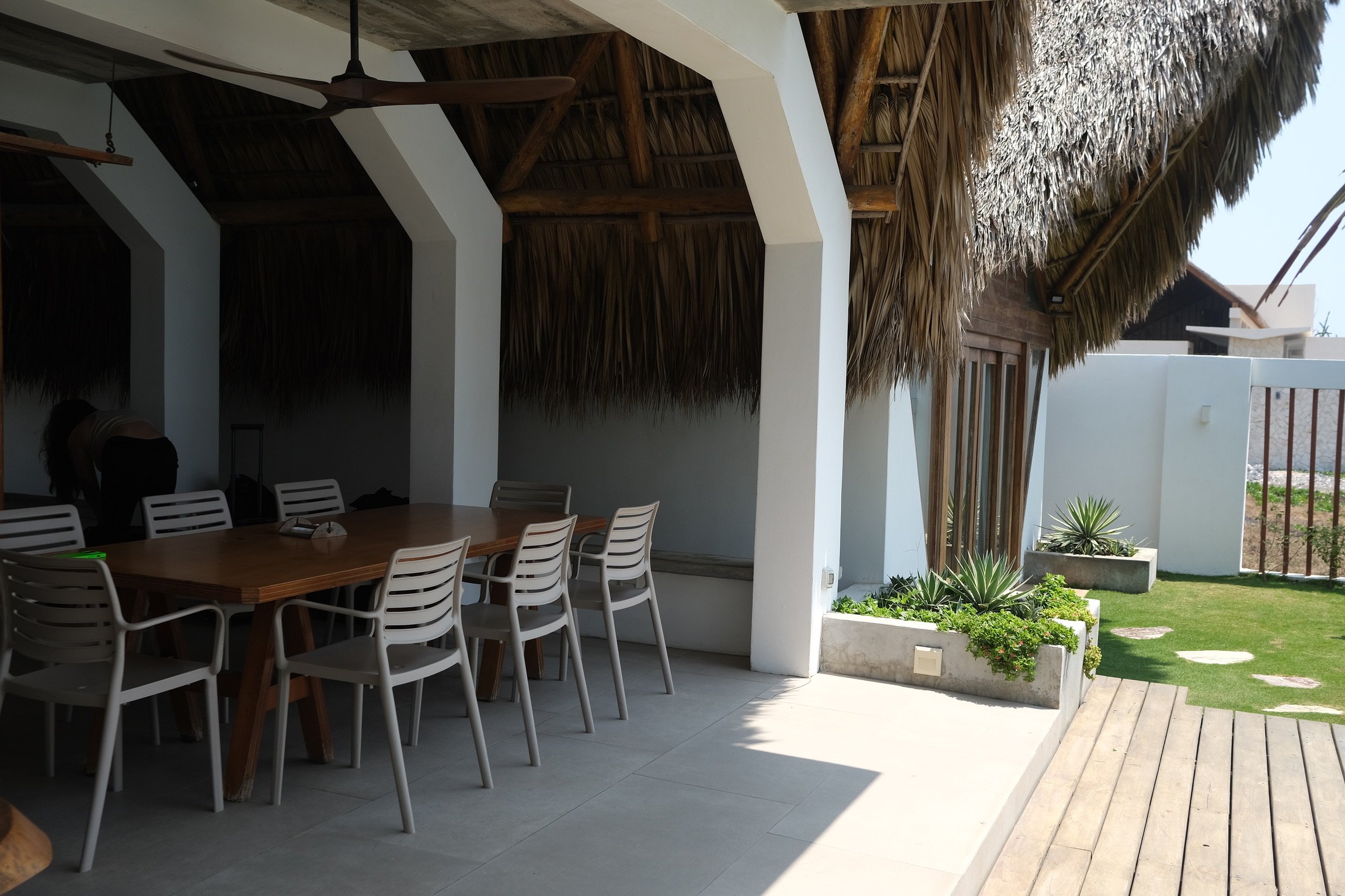 Outdoor patio area with a wooden dining table and white chairs, thatched roof, tropical plants, and a grassy yard with stone stepping stones.