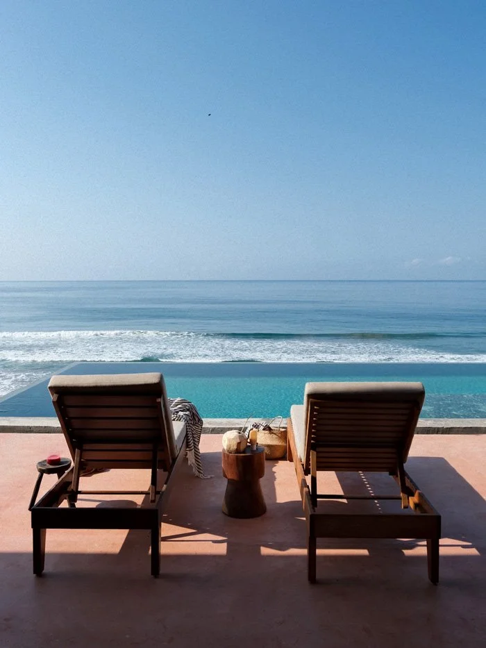 Two lounge chairs on a patio facing the ocean with a small wooden side table between them, pool beside the chairs, and waves in the ocean under a clear blue sky.