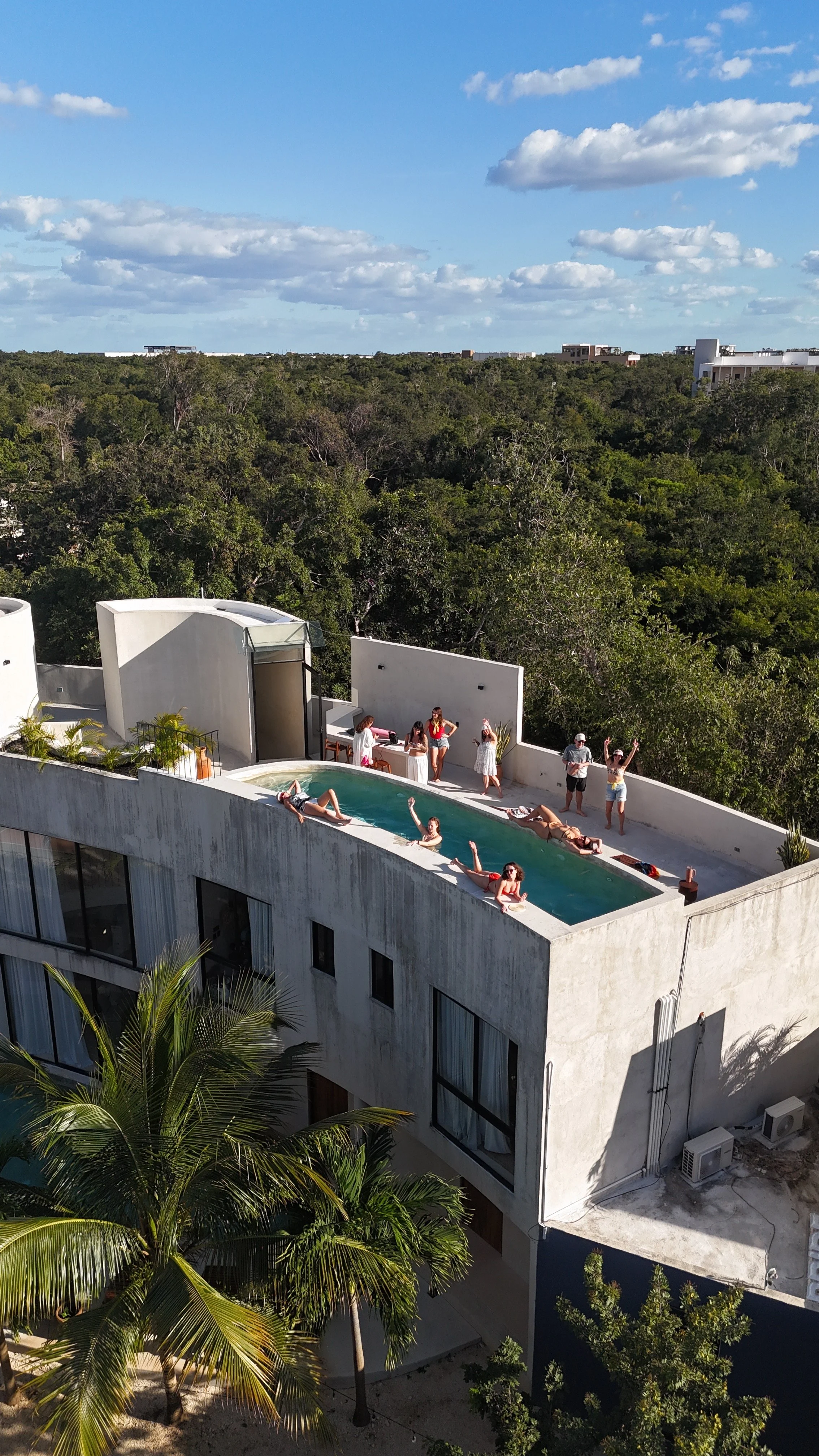 People enjoying a rooftop pool with some sunbathing and socializing, surrounded by trees and cityscape in the distance.