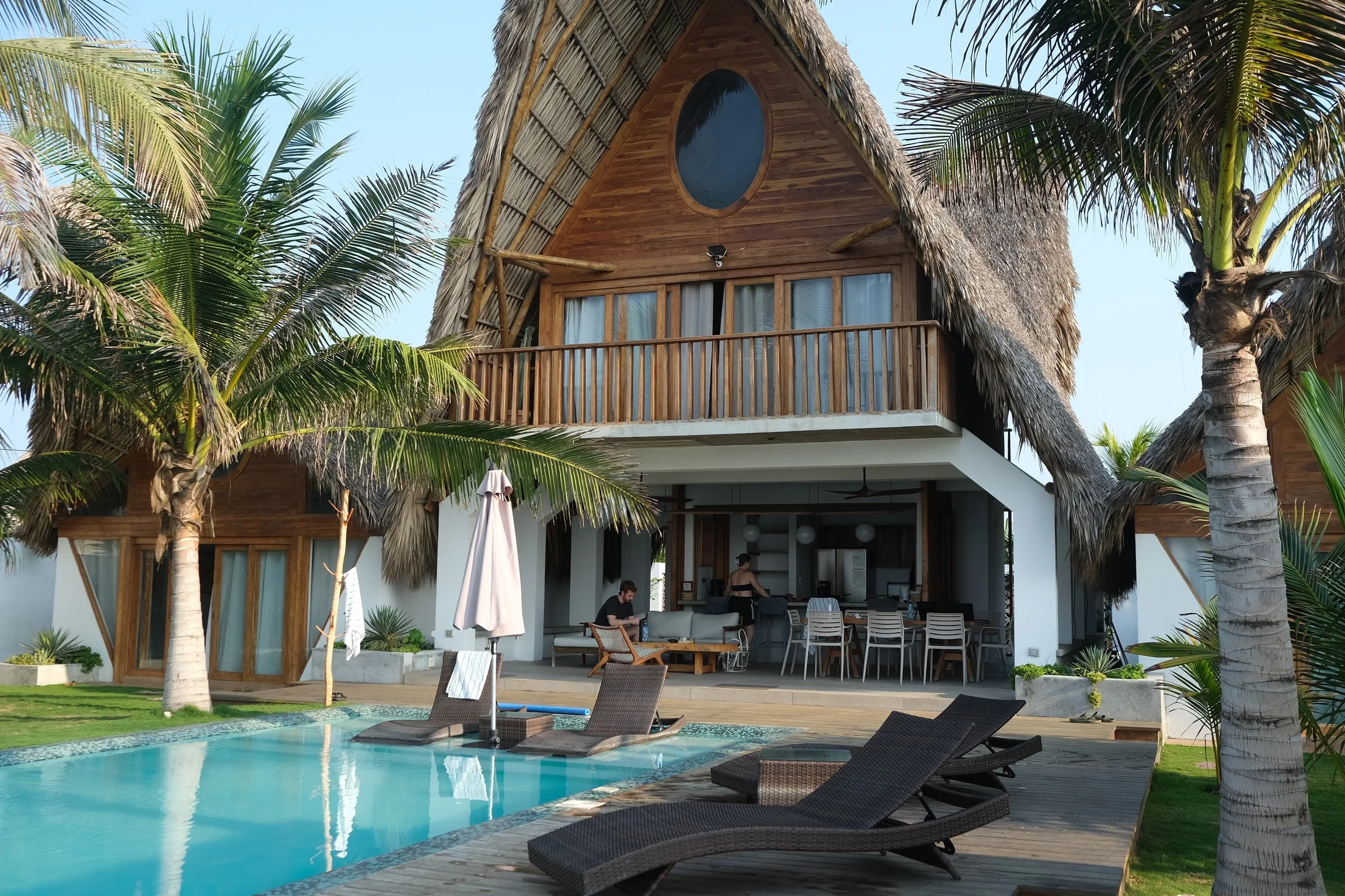 A tropical resort house with a thatched roof, wooden balcony, and large windows, surrounded by palm trees. In the foreground, there is a swimming pool with lounge chairs and umbrellas, with a person sitting by the pool and another preparing inside the house.