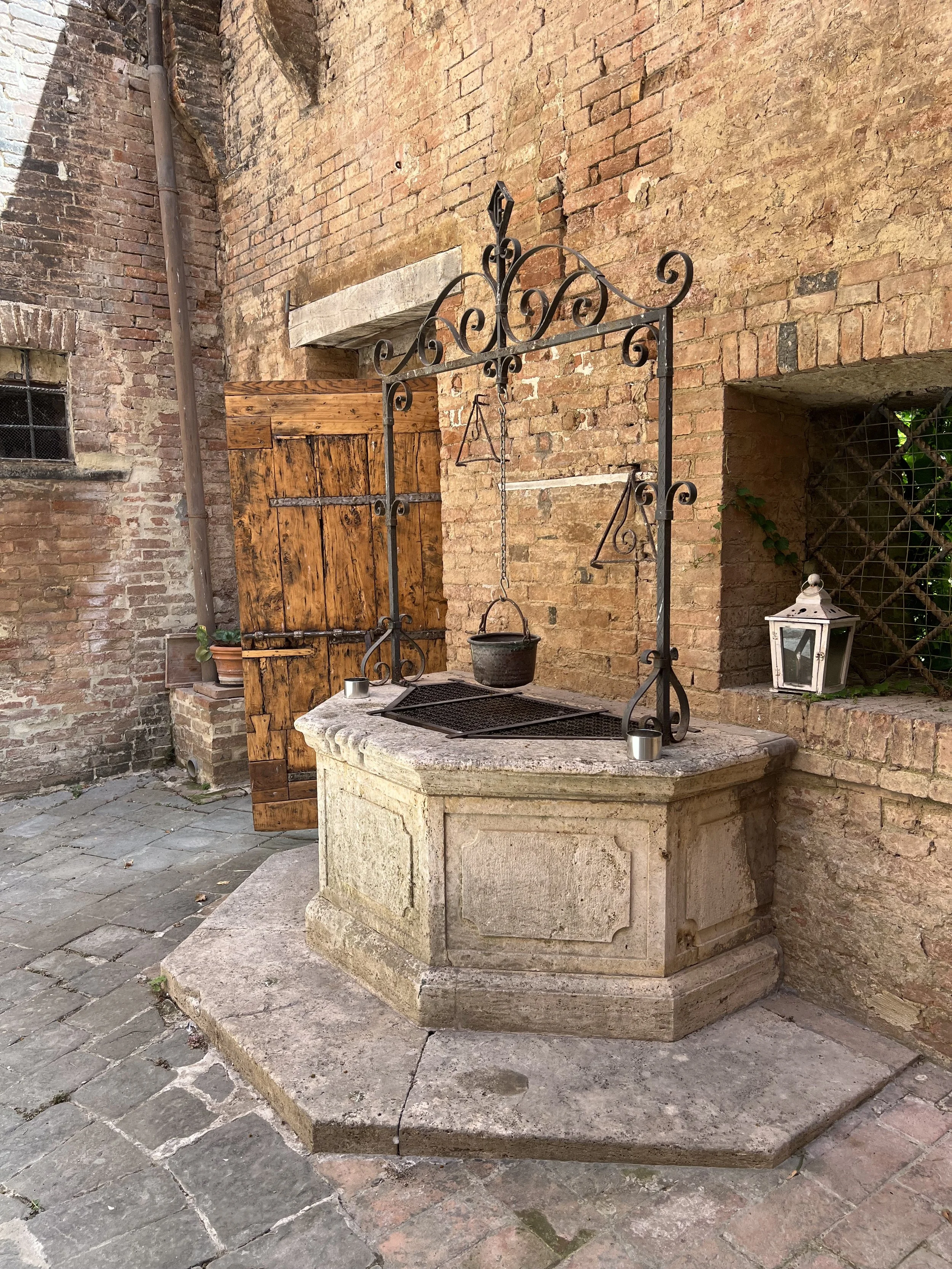 An old stone well with a metal pulley system, a bucket hanging from the pulley, set against a red brick wall in a rustic courtyard.