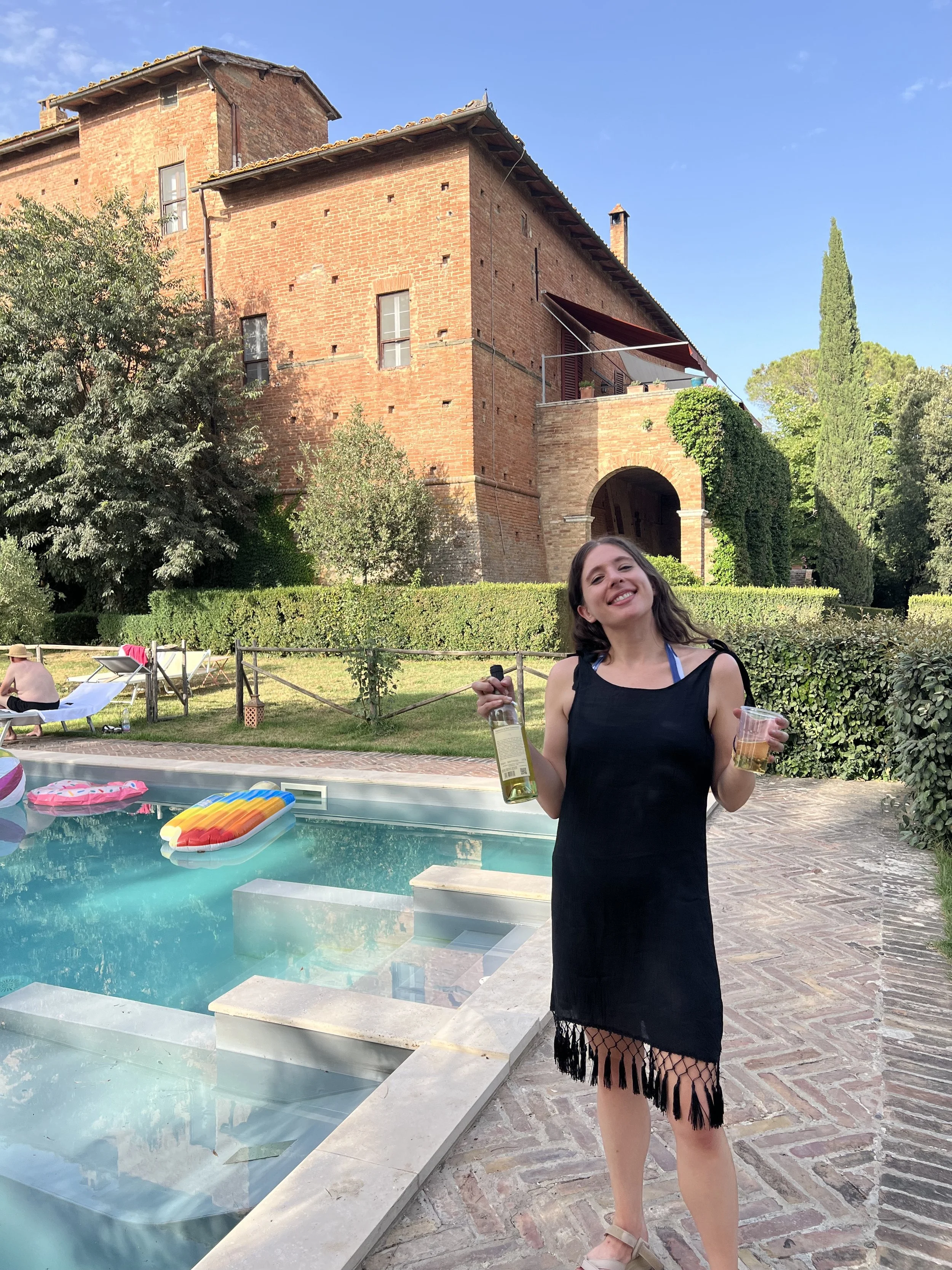 Young woman in a black dress with fringe detail, holding a bottle of white wine and a clear plastic cup with beverage, smiling near a swimming pool with a historic brick building and lush green trees in the background.