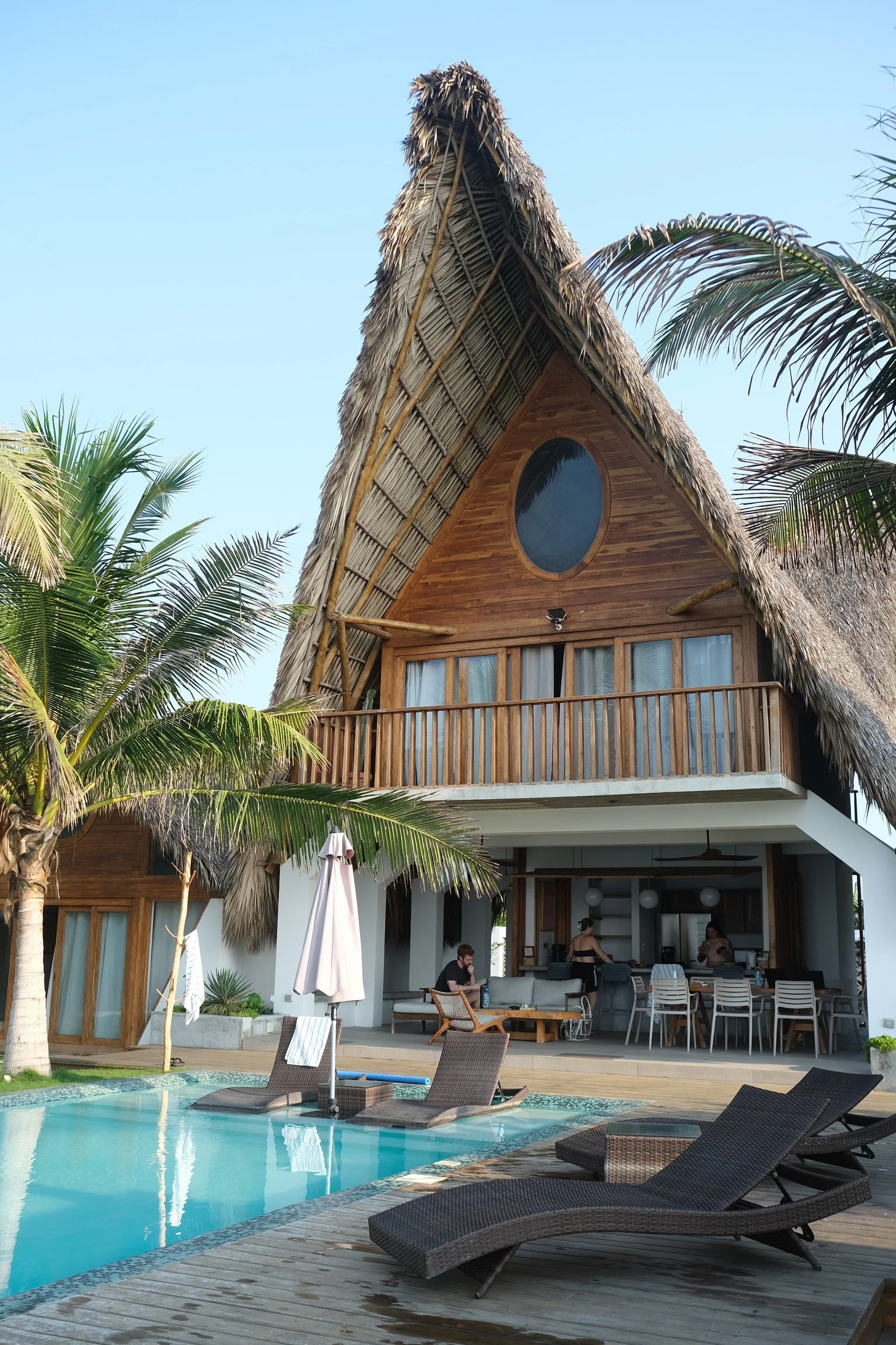 A tropical hotel with a thatched roof, wooden accents, and a pool area with lounge chairs and palm trees.