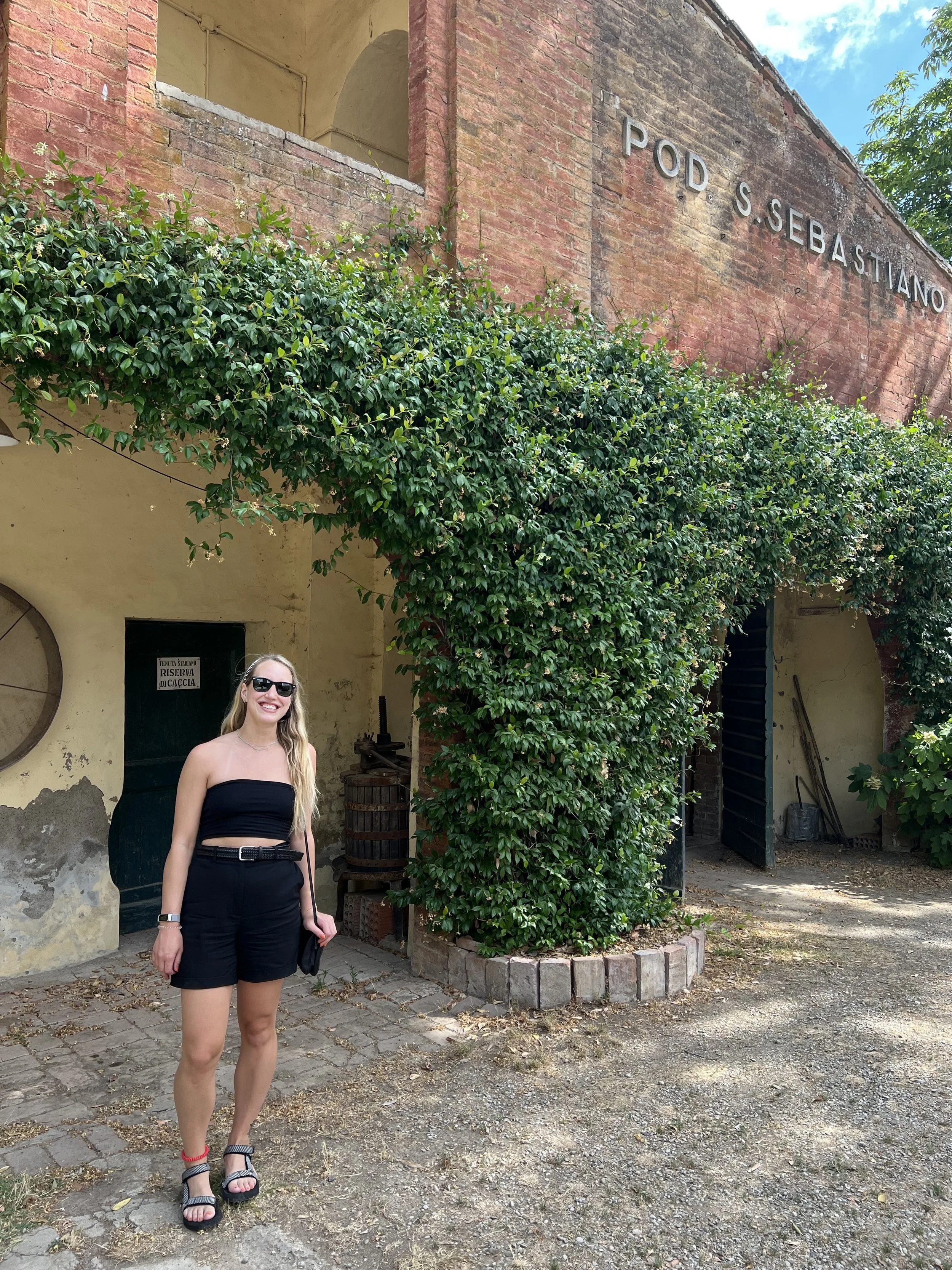 A young woman with long blonde hair, wearing sunglasses, a black strapless top, black shorts, and sandals, stands smiling in front of an old building with a sign that reads 'POD S. SEBASTIANO'. The building has ivy growing over it and a rustic, weathered appearance.