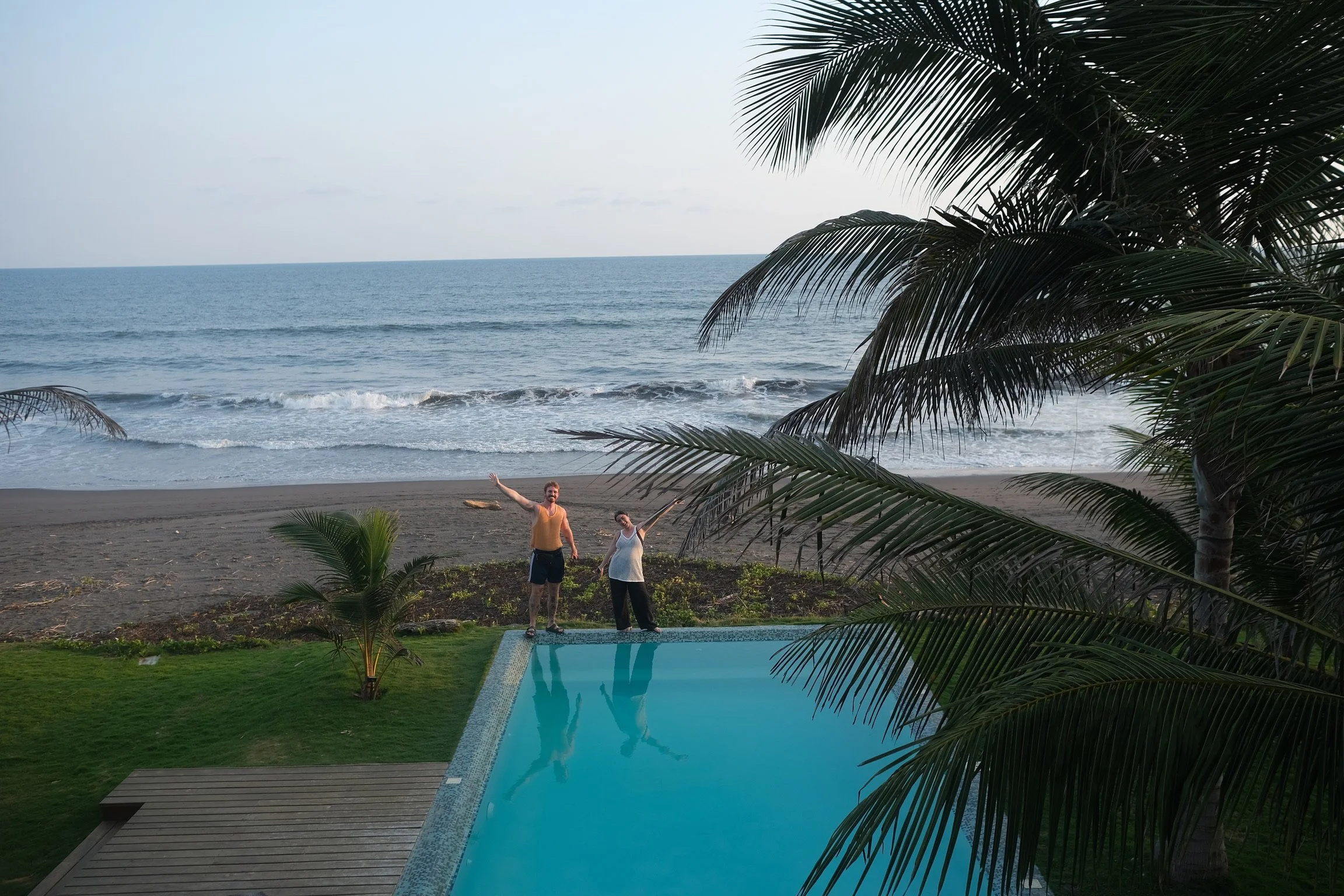 Two people standing next to a swimming pool, with a beach and ocean in the background, surrounded by palm trees.