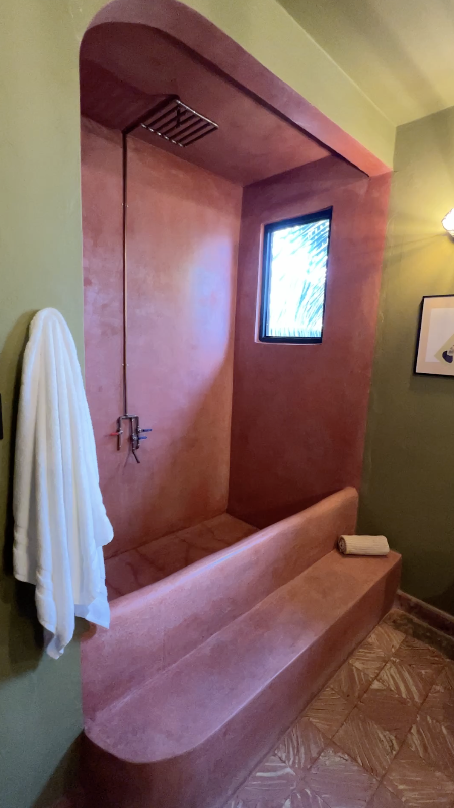 A bathroom with a built-in reddish-brown shower stall, a small window, a white towel hanging on a hook, and a rolled towel on the bench.