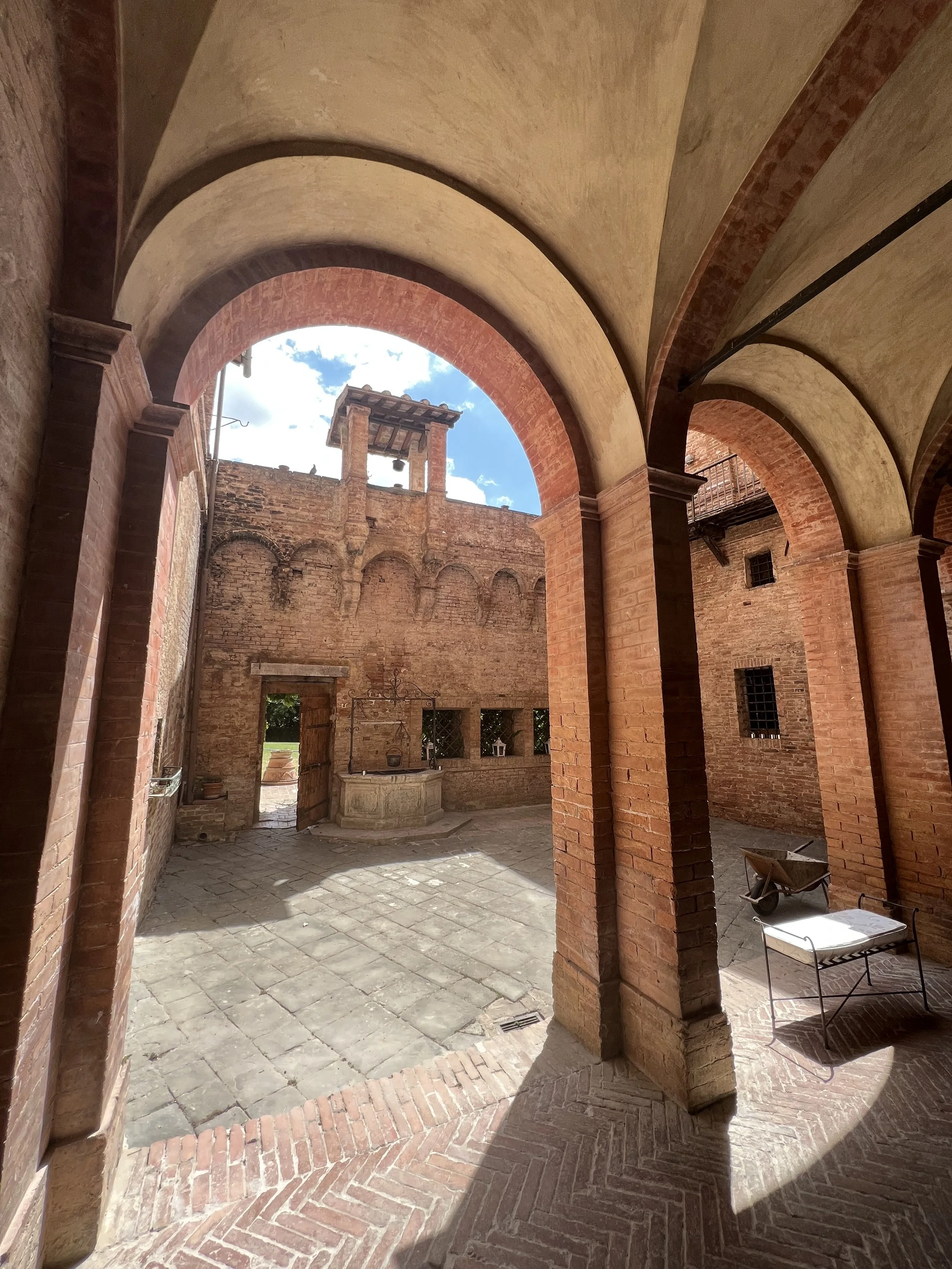 A courtyard with brick walls and arches, a stone fountain, a wheelbarrow, and a bench, with blue sky and clouds overhead.