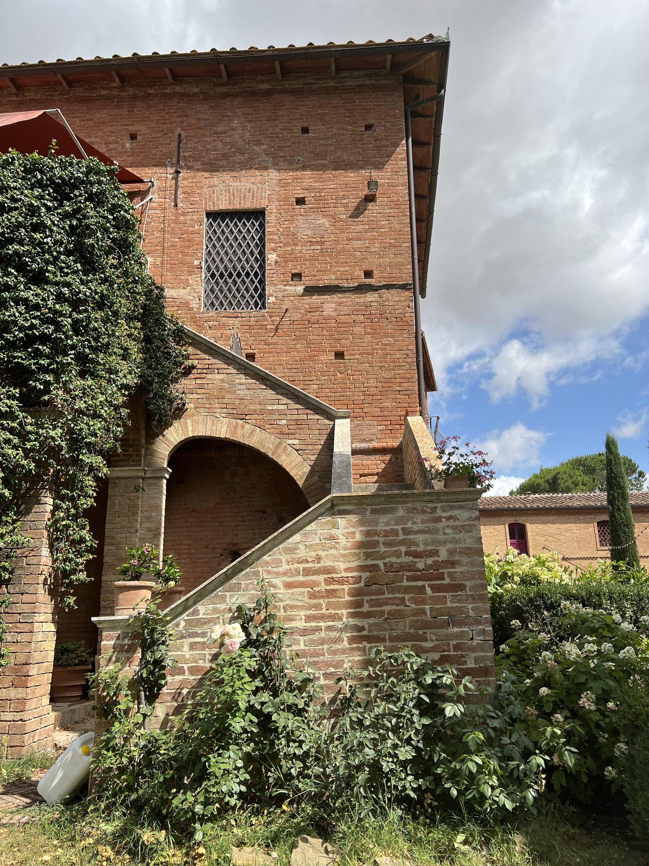 Brick house with arched staircase, surrounded by greenery, under a partly cloudy sky.