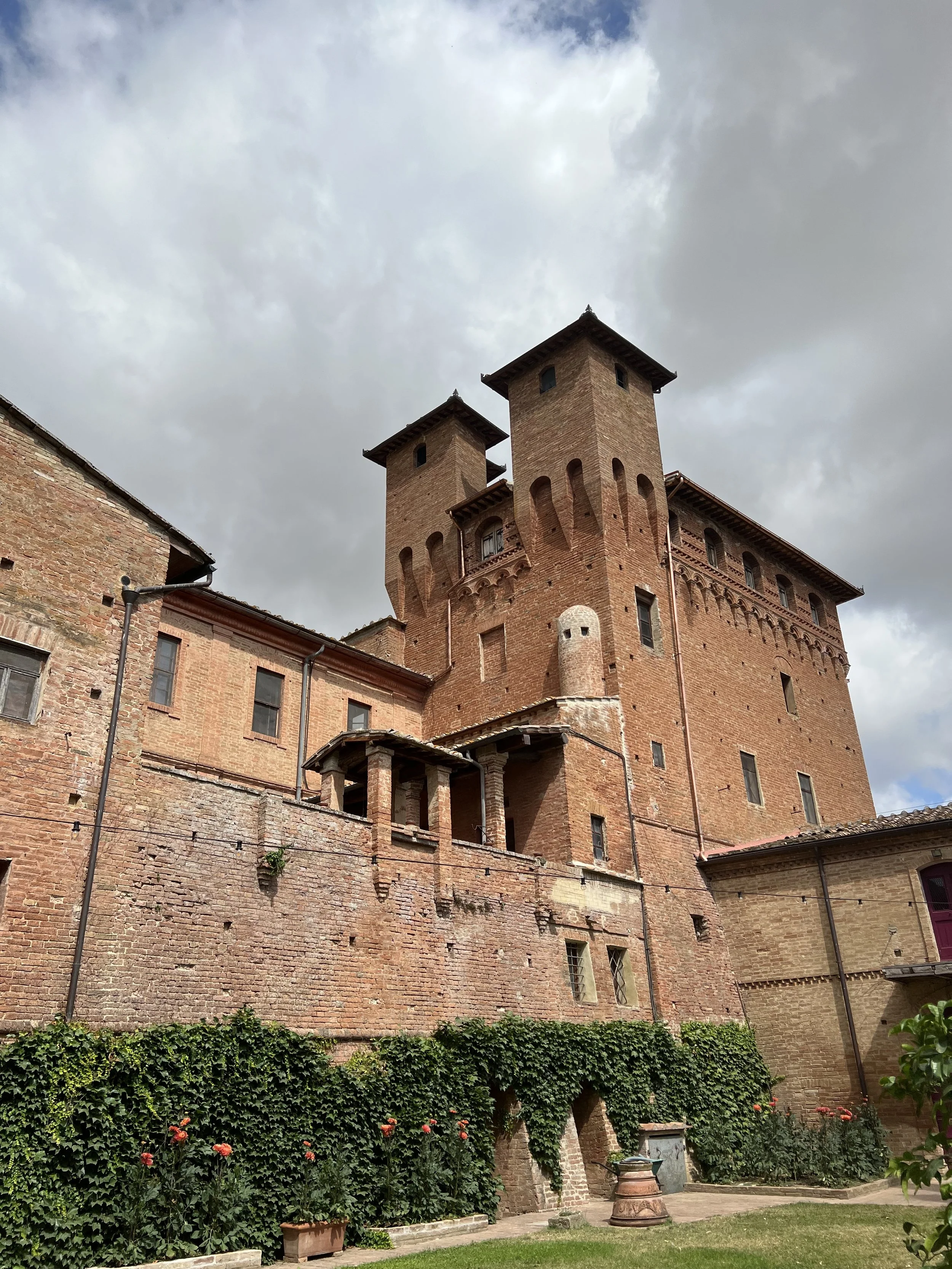 A historic brick castle with multiple towers and small windows, surrounded by greenery and flowers, under a cloudy sky.