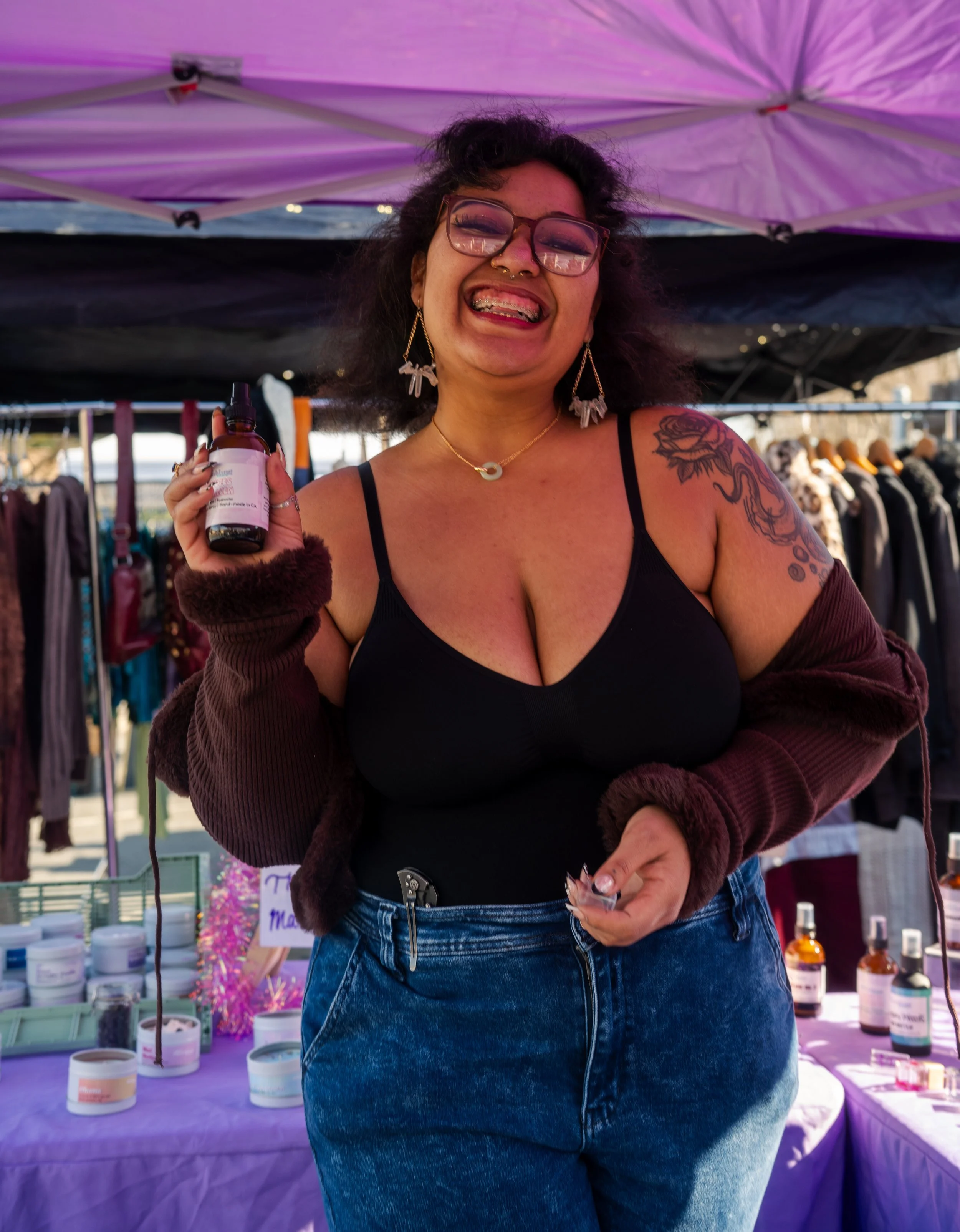 A smiling woman (Olivia Williams, Owner of Many Mumbling Mice) with tattoos on her arms, lying on a patterned blanket surrounded by candles, disco balls, and decorative stones, making a playful face with her 3-in-1 Room Spray in her right hand.