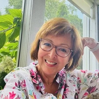 Woman with glasses wearing a floral blouse, smiling indoors with greenery in the background.