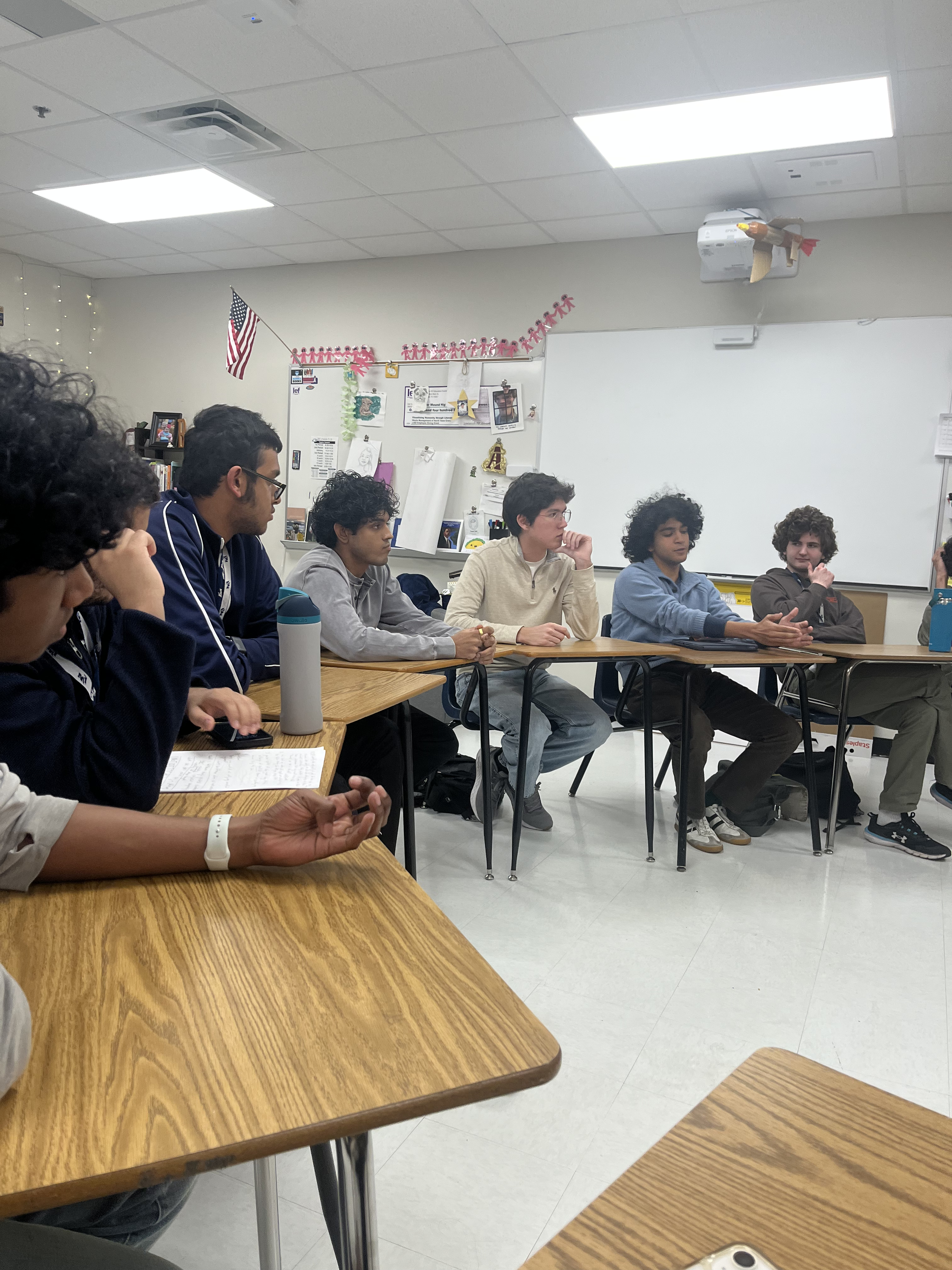 A classroom with seven students sitting at desks, engaged in a discussion or listening to a speaker, with a whiteboard and classroom decorations in the background.