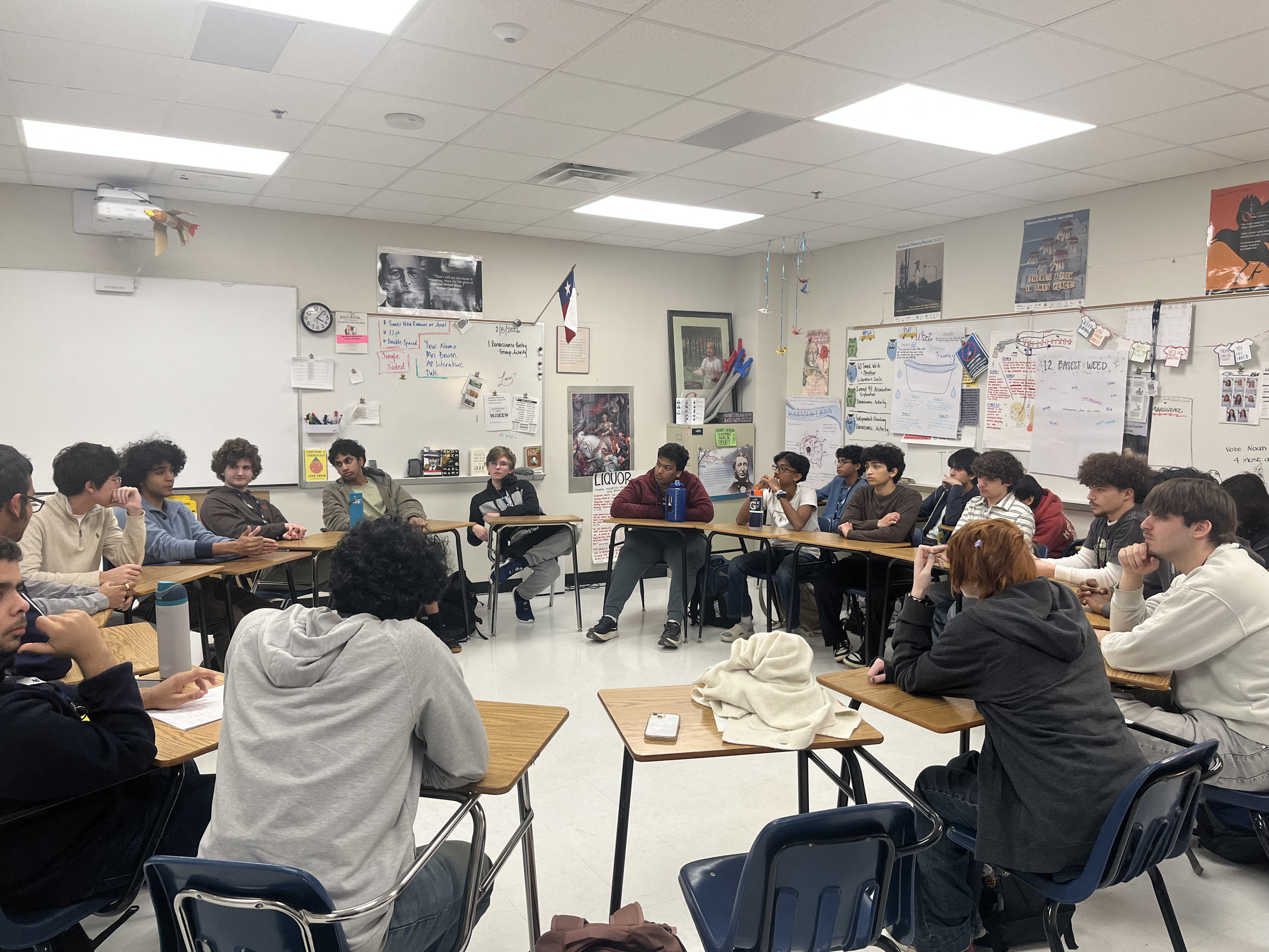 A classroom filled with students seated around tables forming a U-shape, listening to a teacher or speaker not visible in the image. The room has white walls with various posters, notes, and a whiteboard filled with writings and images.