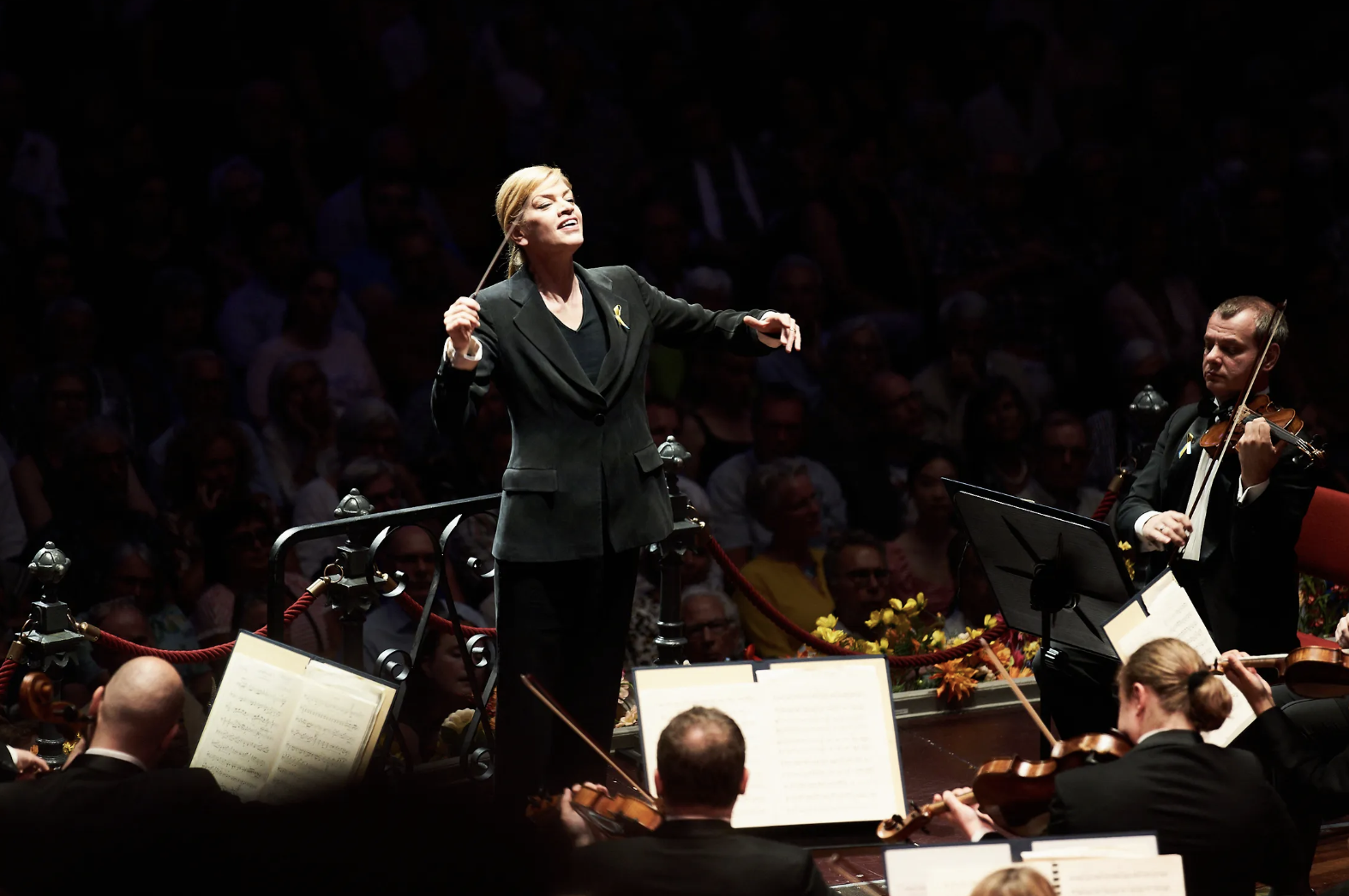 Conductor Keri-Lynn Wilson leading the Ukrainian Freedom Orchestra during a performance, with musicians playing violins and sheet music visible, and an audience watching in the background.
