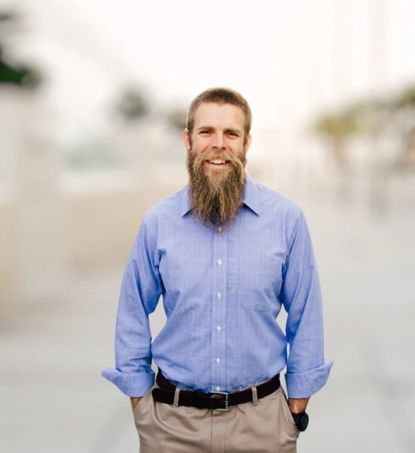 Smiling man with a beard, wearing a blue shirt and khaki pants, standing outside with a blurred background.