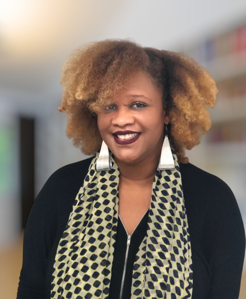 Smiling person with curly hair wearing a patterned scarf and black top, standing indoors in a softly blurred background.