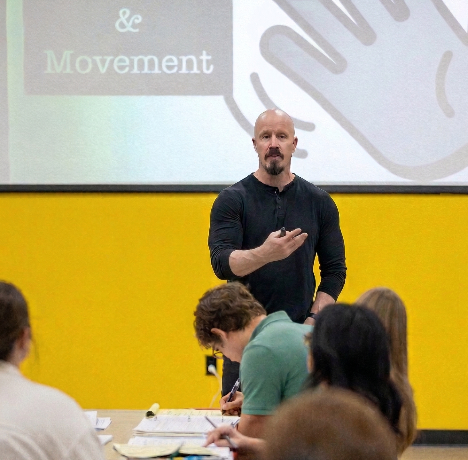 A man with a goatee and bald head, wearing a black long-sleeve shirt, standing in front of a yellow wall, giving a presentation to an audience. A large screen behind him displays the words '& Movement' with a graphic of a hand.