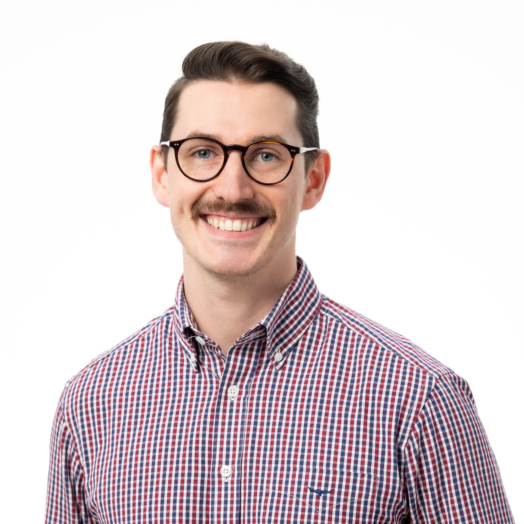 A smiling man with dark hair and glasses wearing a checkered shirt against a white background.