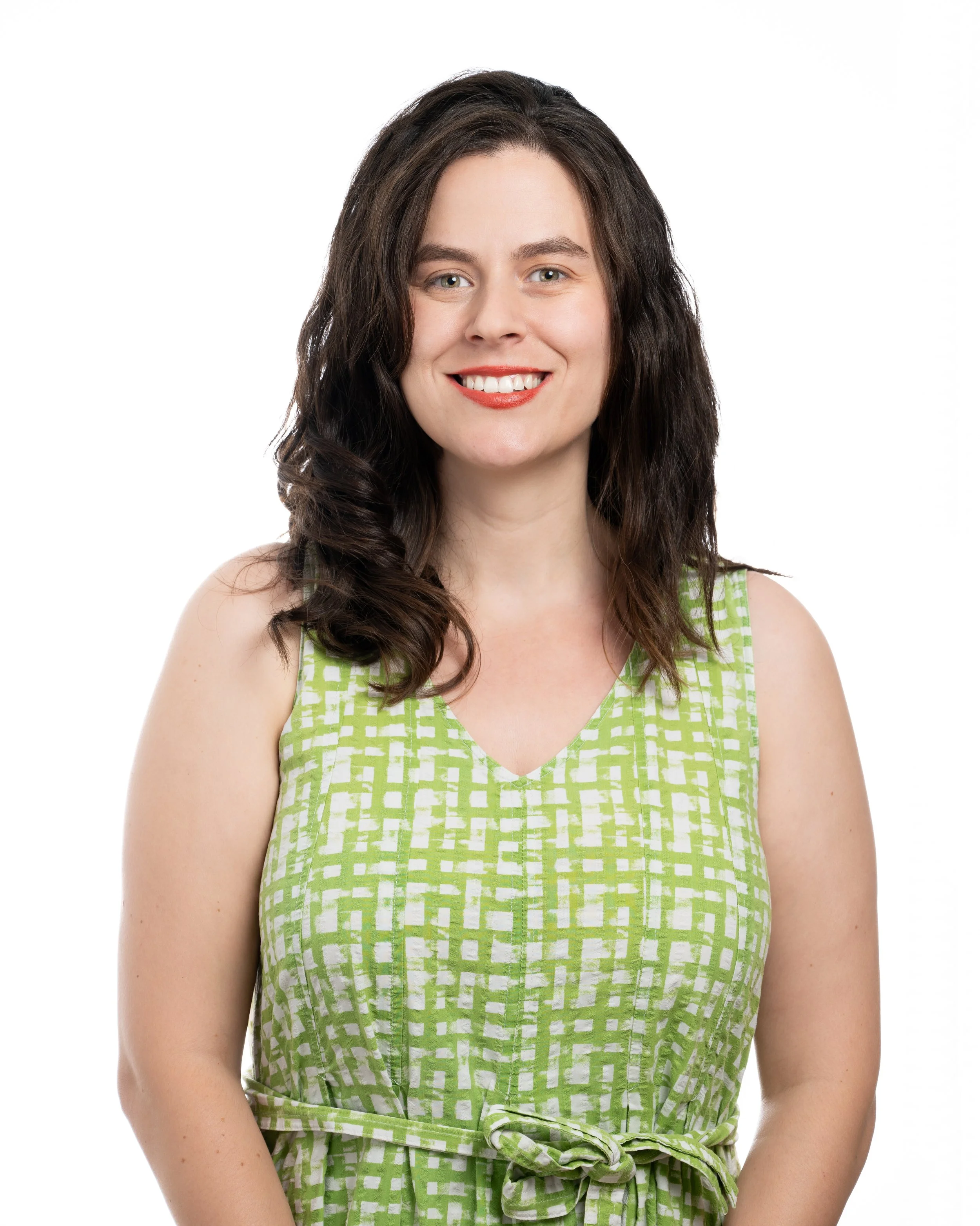 A woman with shoulder-length dark brown hair, wearing a sleeveless green and white patterned dress, smiling at the camera against a plain white background.