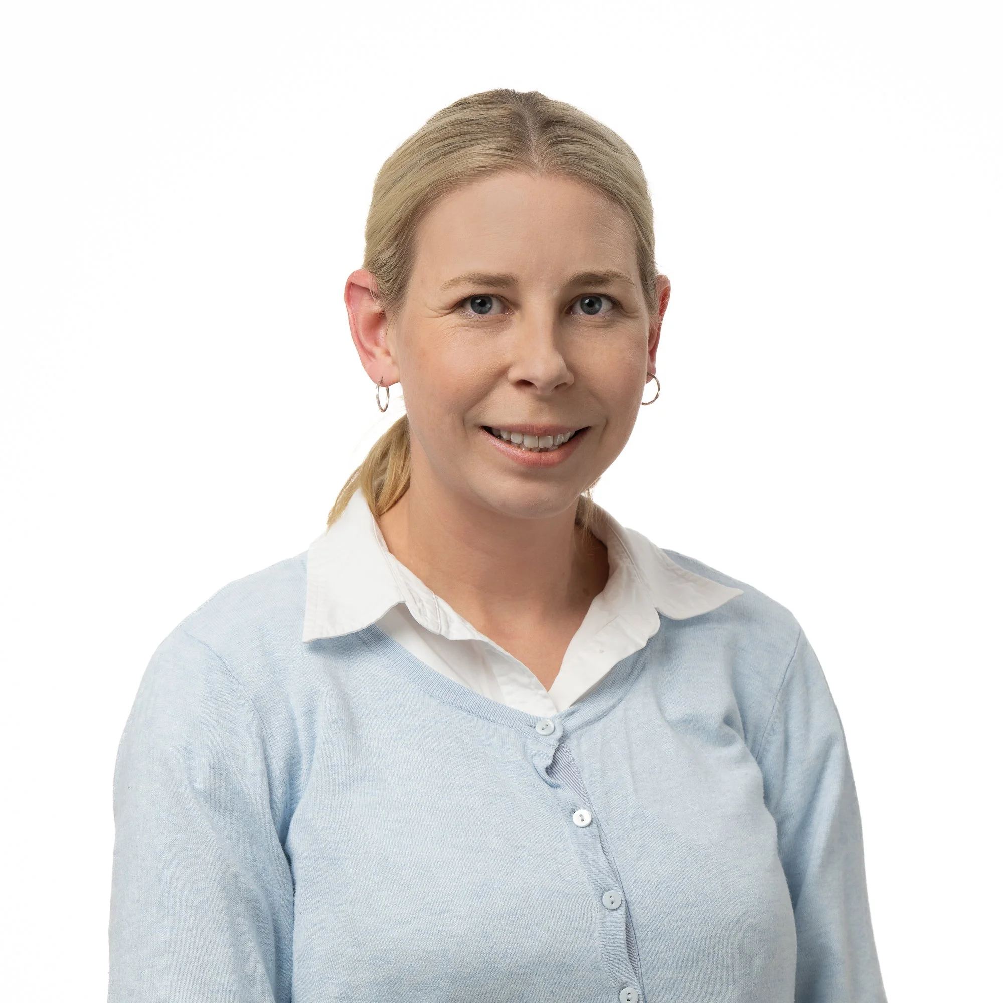 A young woman with blonde hair tied back, wearing a white collared shirt under a light blue cardigan, smiling at the camera against a white background.