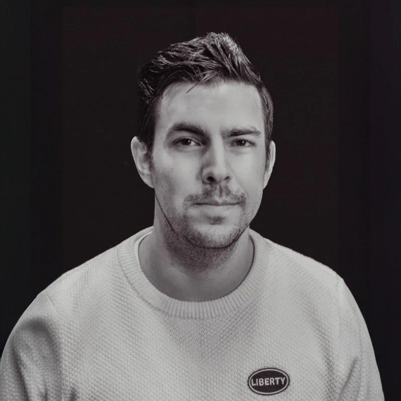 A young man with styled dark hair and a slight beard, wearing a light-colored sweater with a Liberty logo, smiling slightly against a dark background.