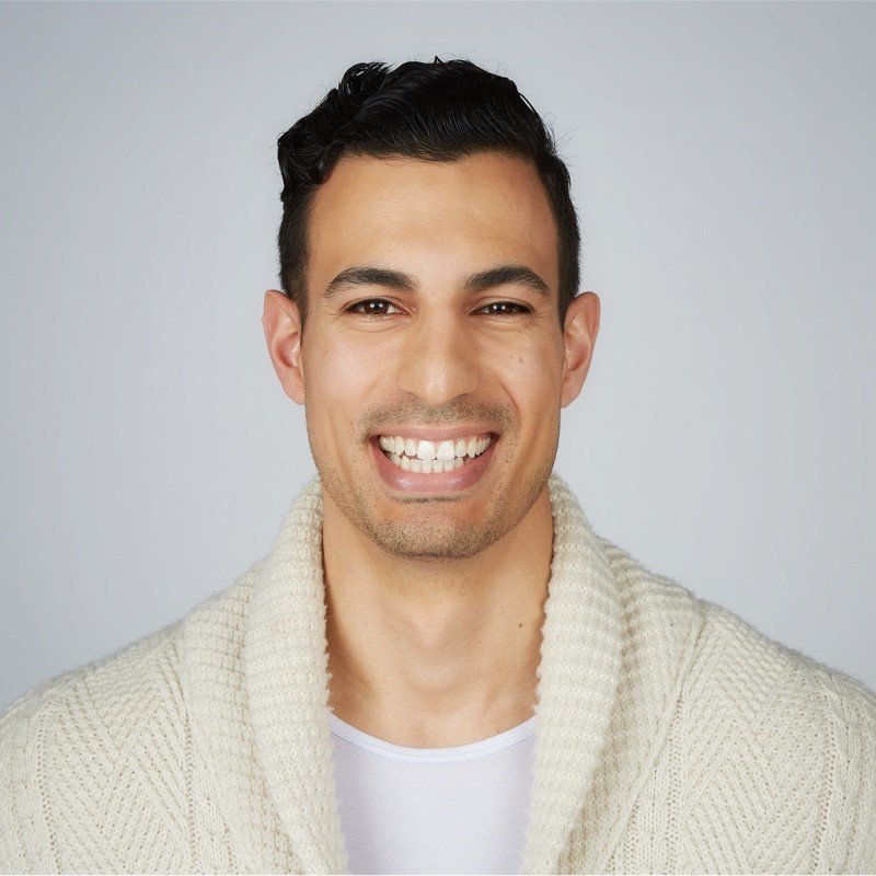 Young man with dark hair smiling, wearing a cream-colored sweater against a light gray background.