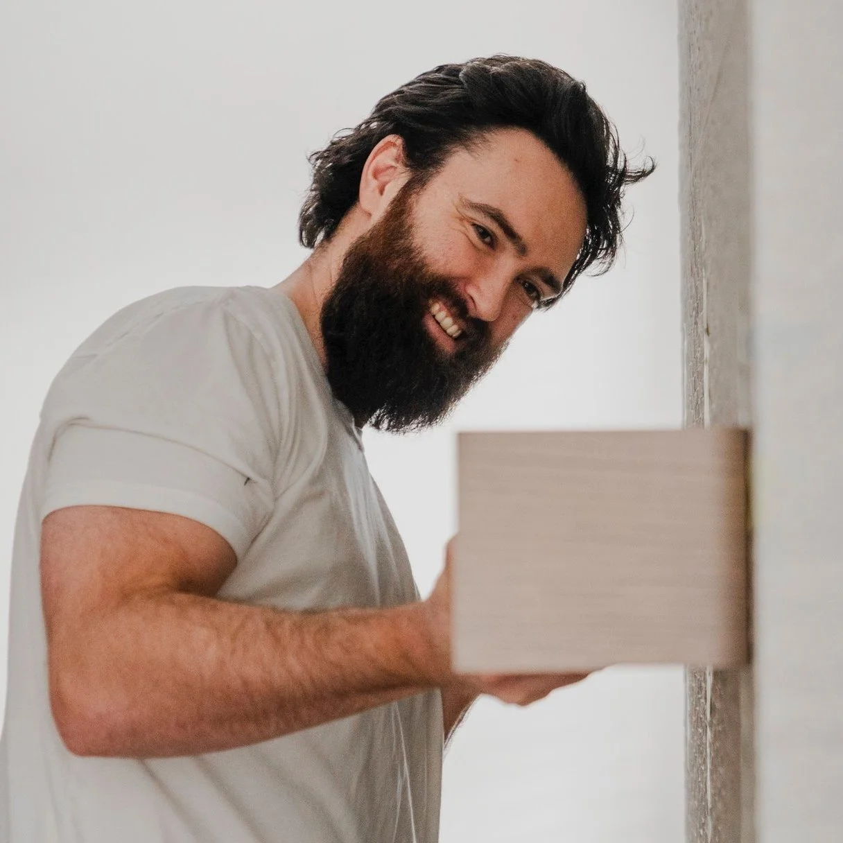 A smiling man with a beard and long hair, wearing a white t-shirt, holds a stack of white tiles as he looks at the camera.