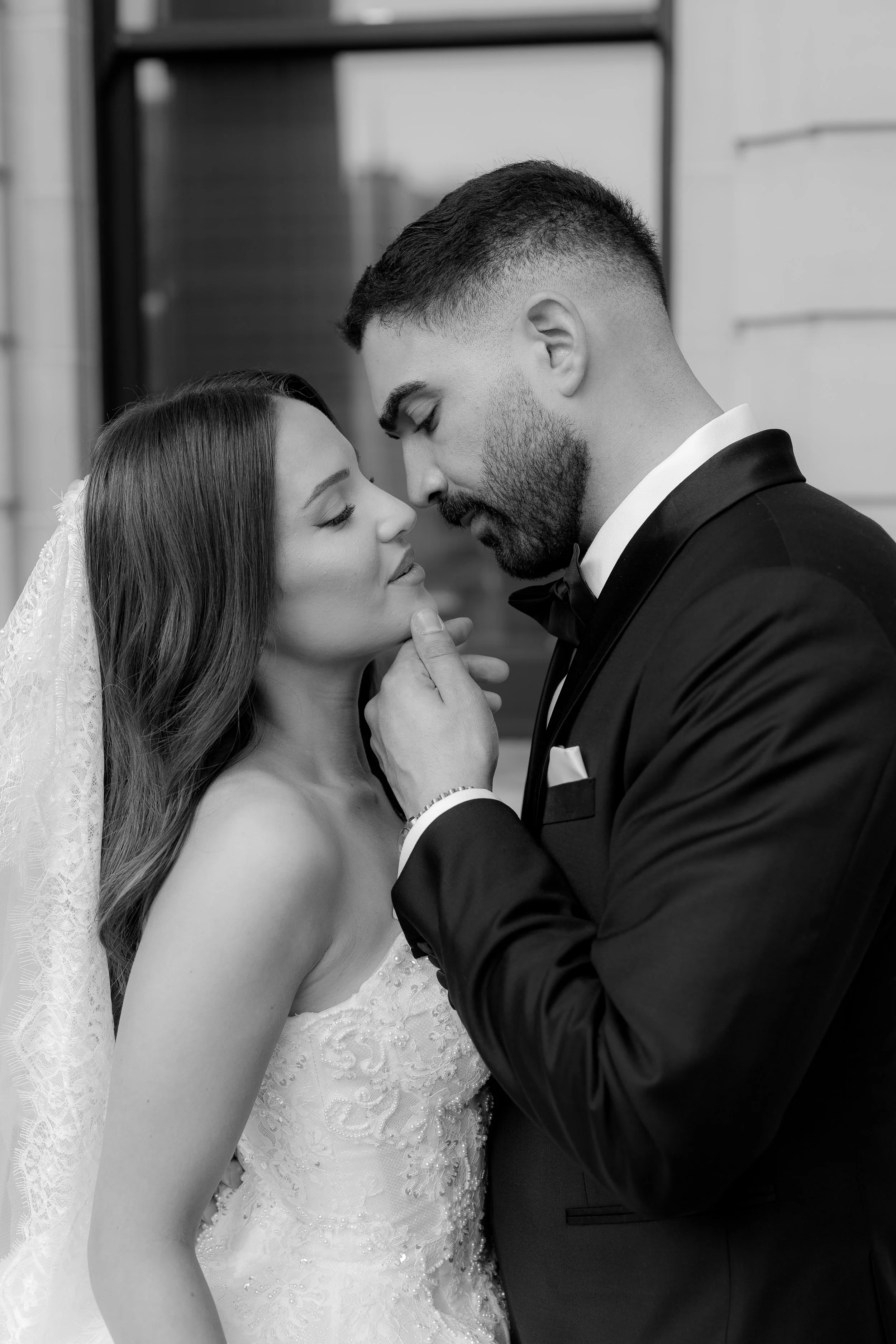 A bride and groom in wedding attire sharing an intimate moment, with their faces close and eyes closed, outside of a building.