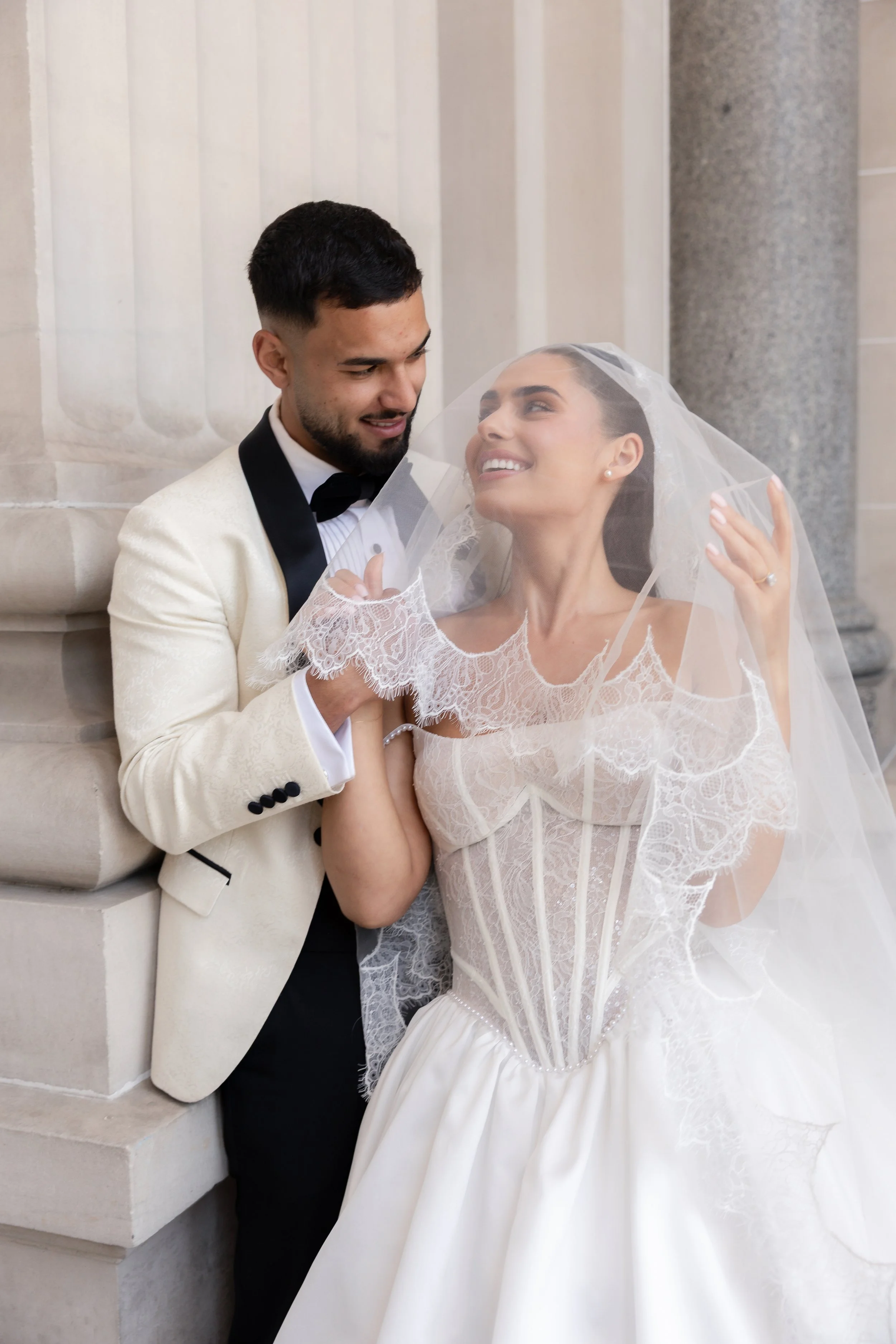 A bride and groom sharing a joyful moment on their wedding day outside a building. The bride is wearing a white wedding dress with lace details and a veil, and the groom is dressed in a cream tuxedo with black lapels and a black bow tie. The bride is
