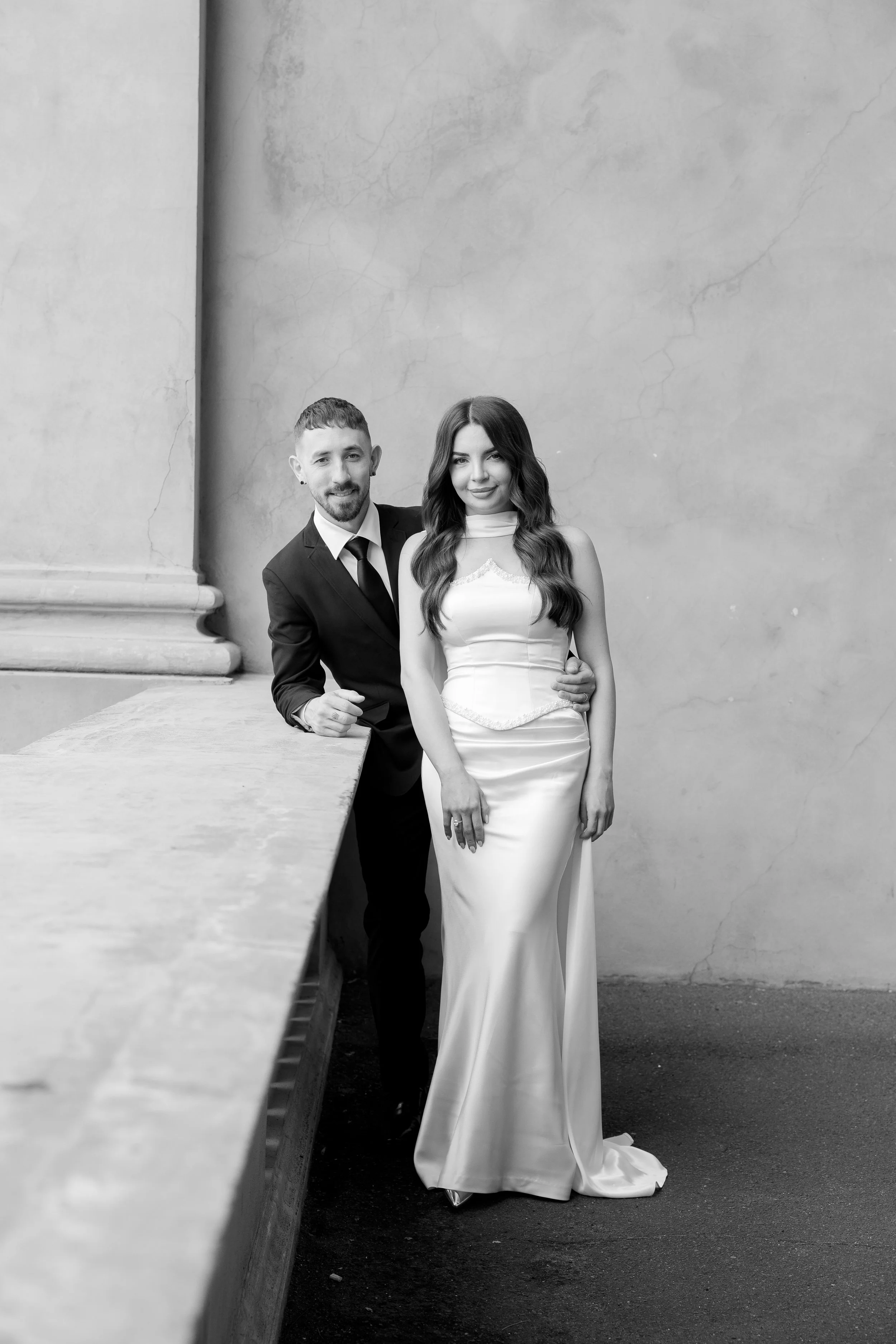 A black and white photo of a man in a suit and a woman in a wedding dress, standing outdoors against a concrete wall. The woman has long, wavy hair and the man has short hair and a beard.