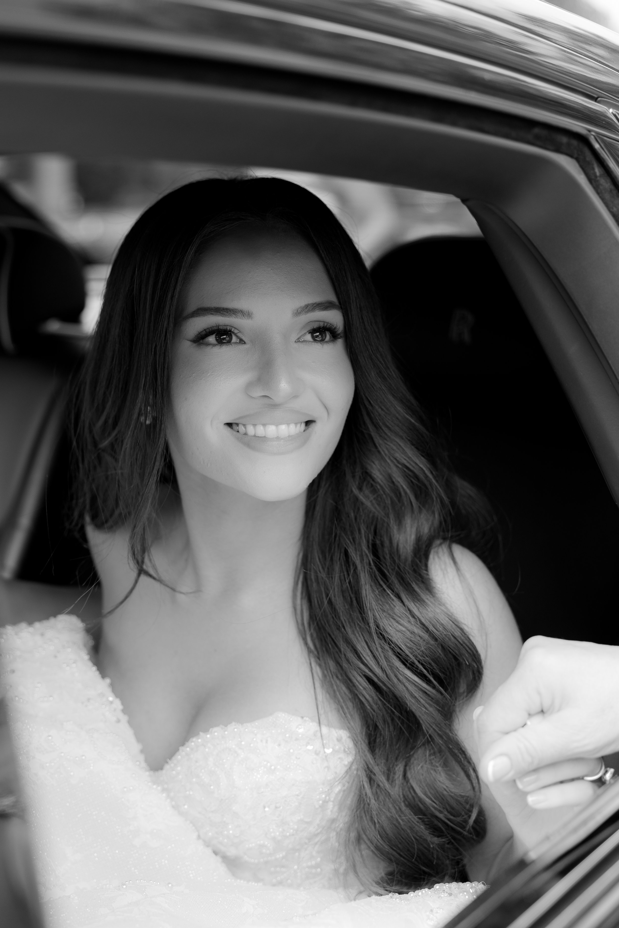 A smiling woman with long dark hair, in a wedding dress, sitting in a car and holding hands with someone outside the car.