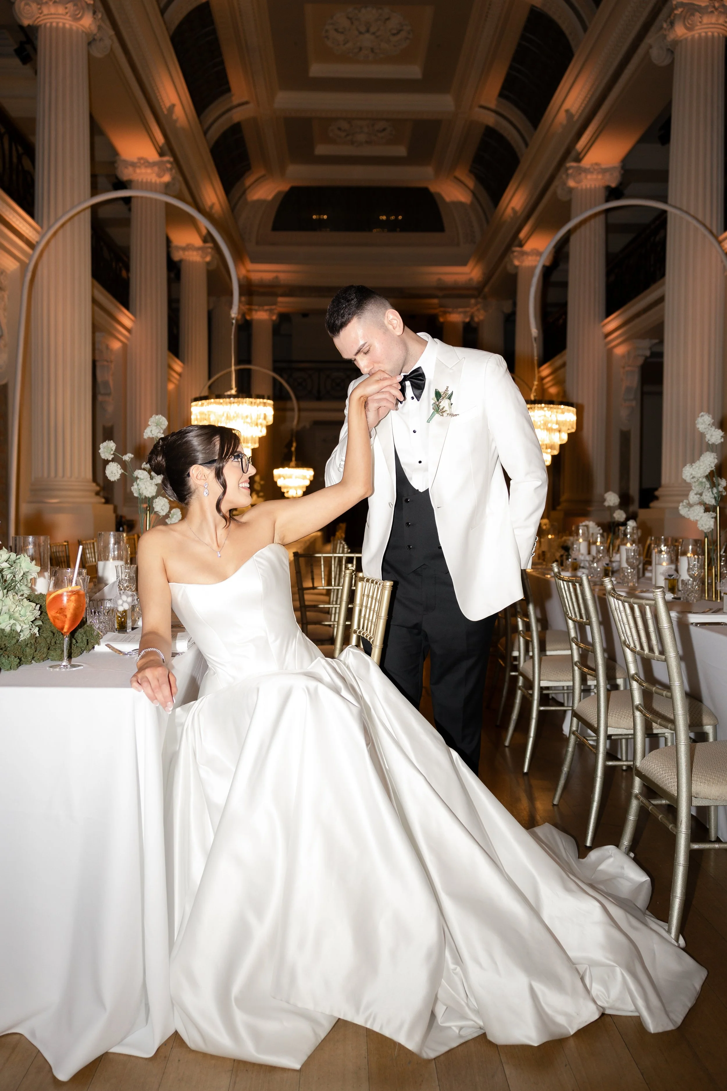 A bride and groom at their wedding reception. The bride is sitting at the table, smiling and touching the groom's chin, who is standing and leaning toward her, dressed in formal wedding attire. The setting is an elegant banquet hall with chandeliers,