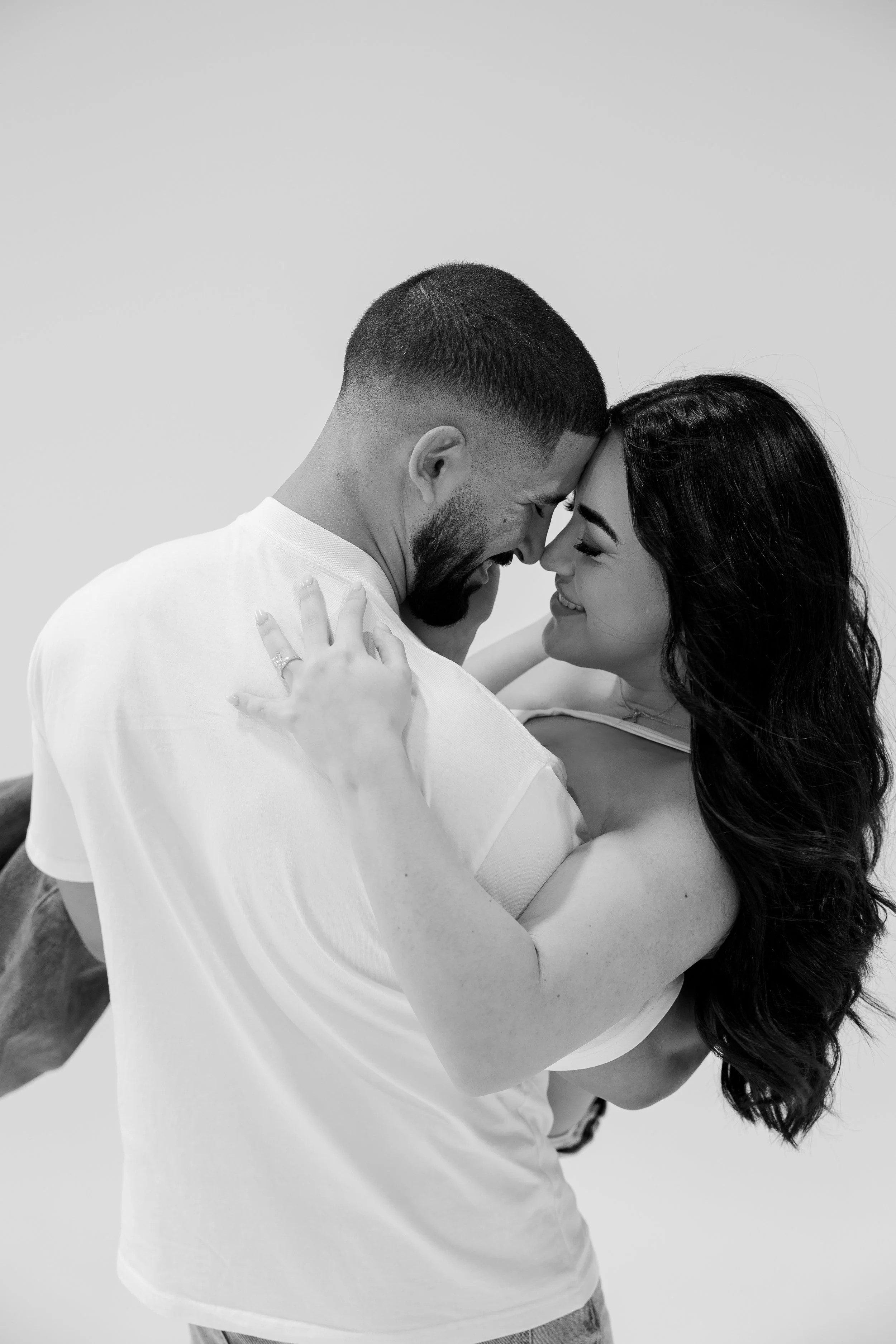 A couple sharing a close, joyful moment, face-to-face, in a black-and-white photo. The man holds the woman, who has long dark hair, as they touch foreheads and smile at each other.