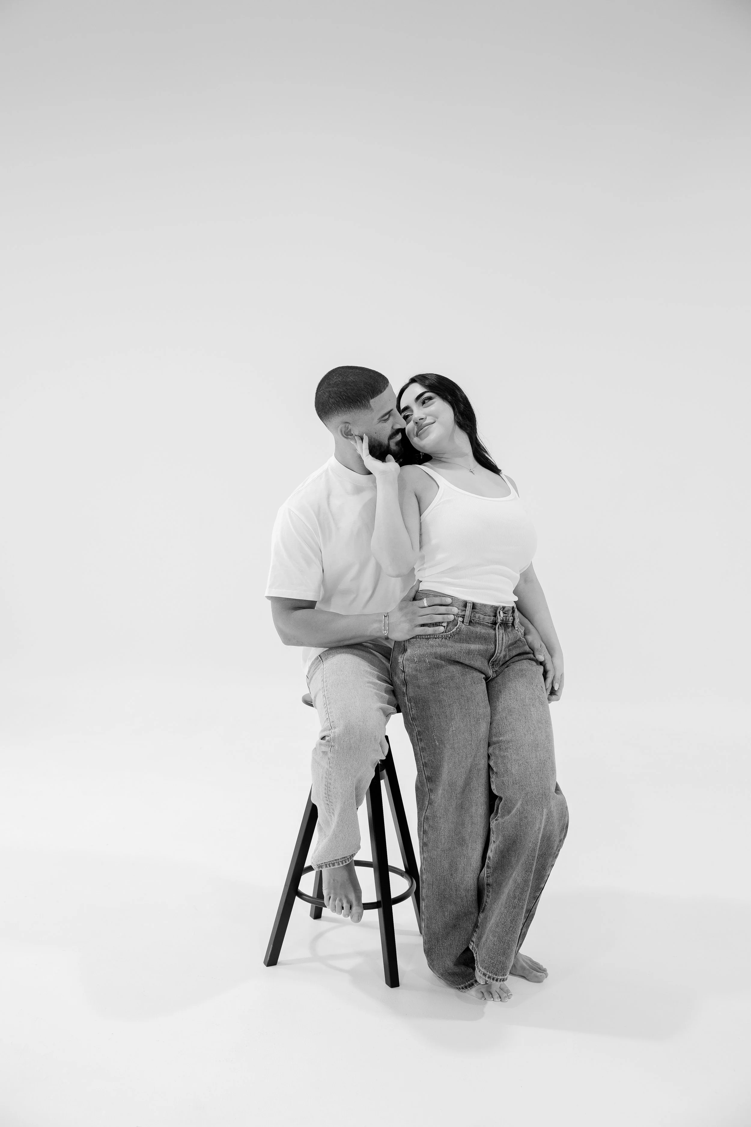Black and white photo of a man sitting on a stool and woman standing beside him, touching her face and smiling at each other.