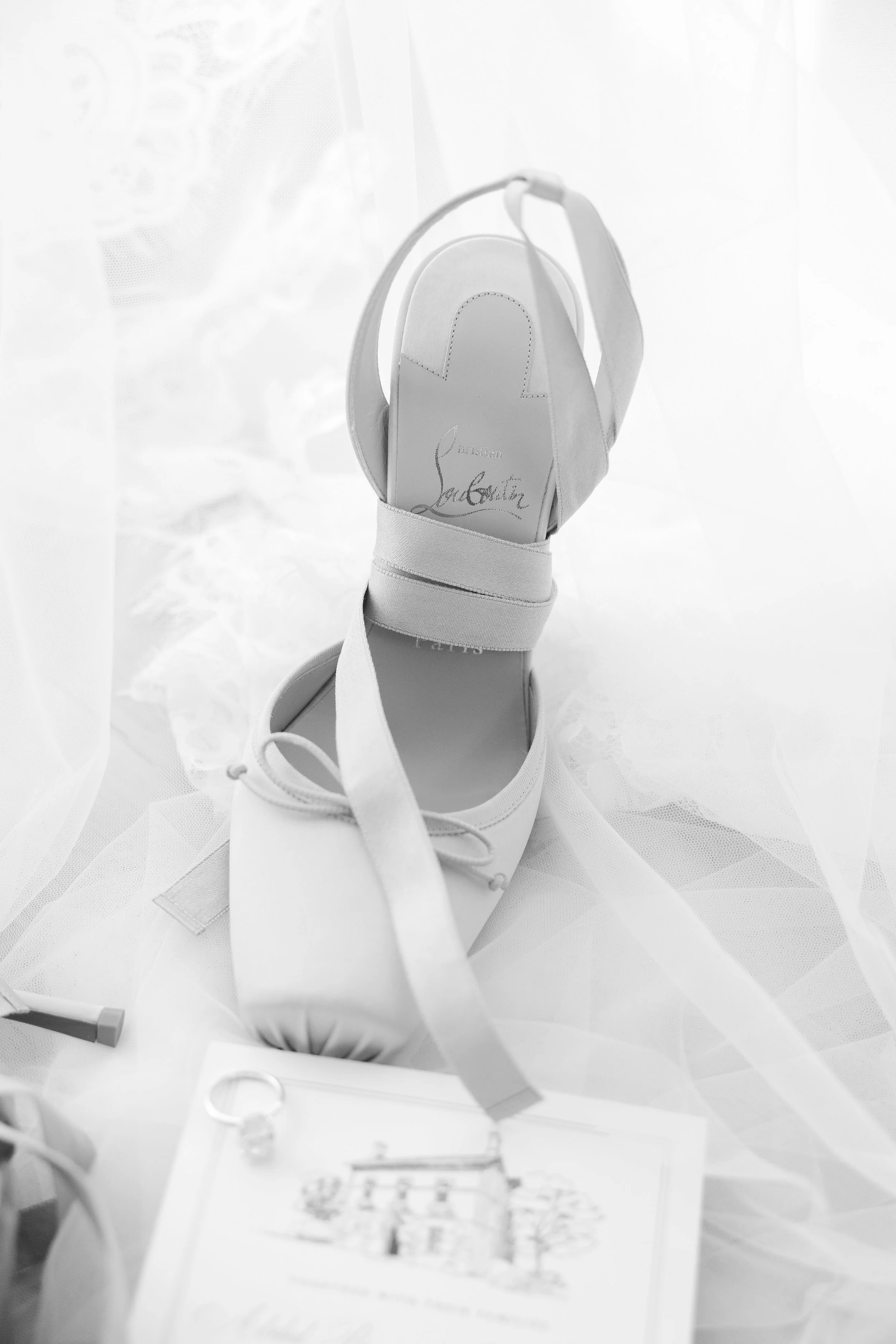 A white designer high heel shoe with a strap and a bow detail, placed on a soft fabric surface, with a white picture frame and small decorative items nearby.