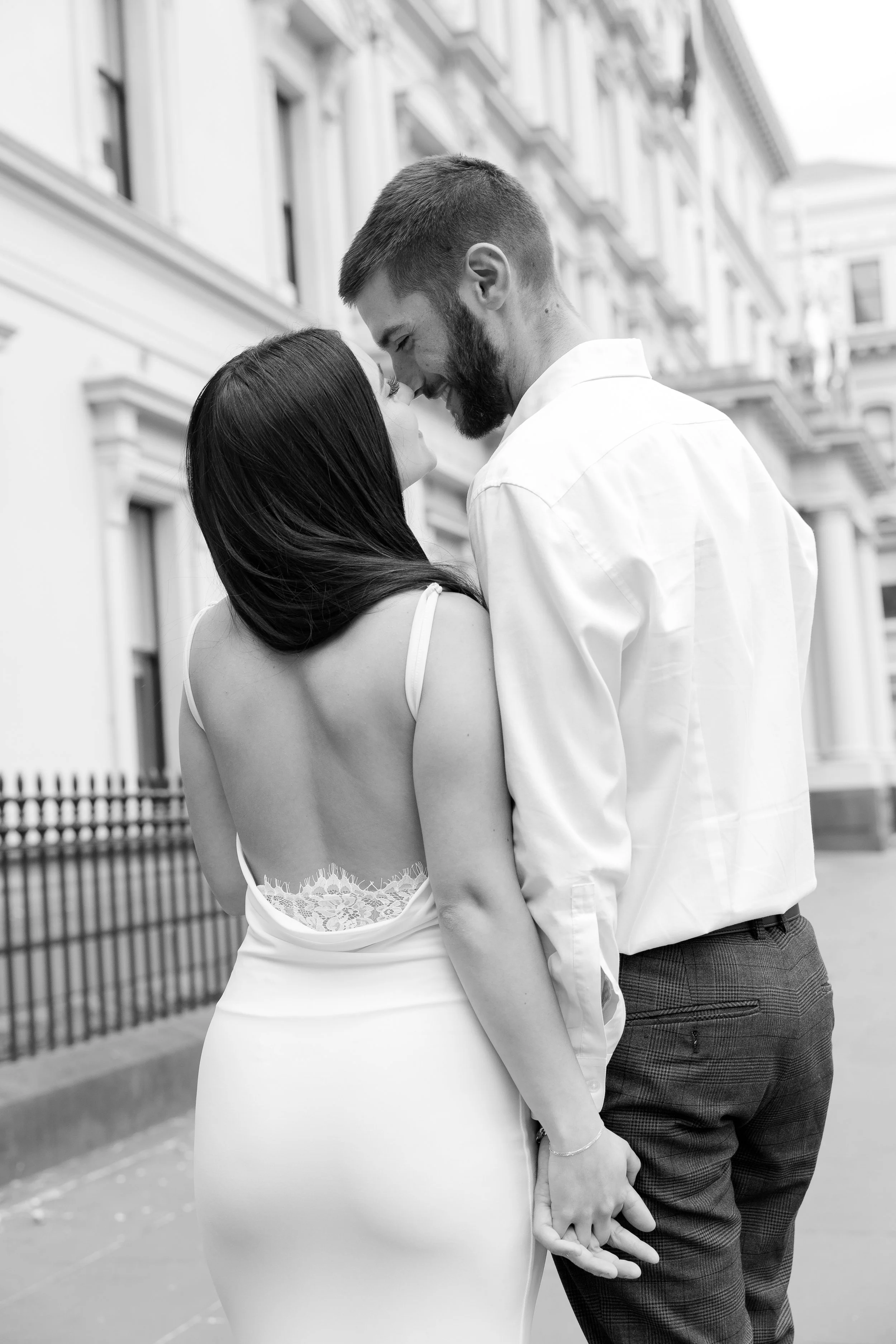 A couple sharing an intimate moment outdoors in front of a row of city buildings, holding hands and leaning their foreheads together.