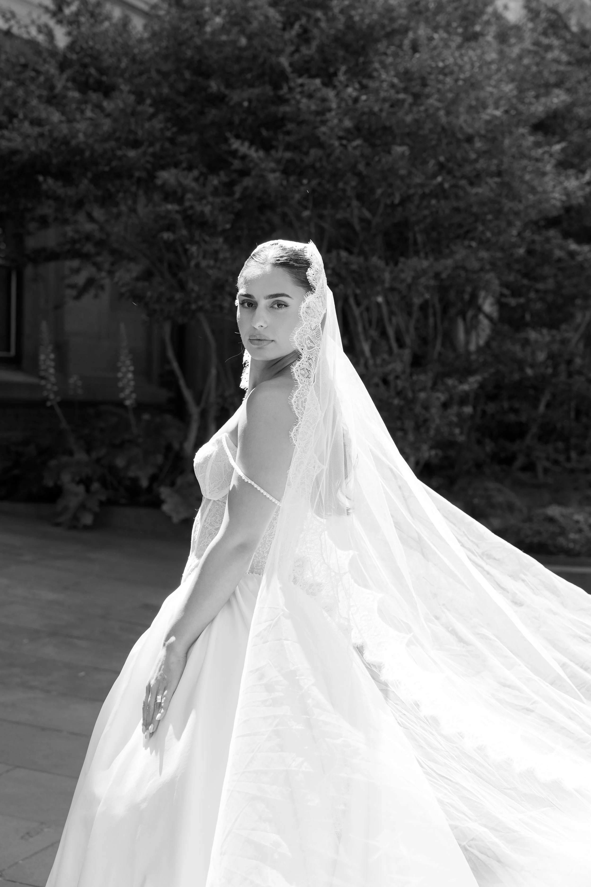 Young woman in wedding dress with veil standing outdoors, with trees and a building in the background.