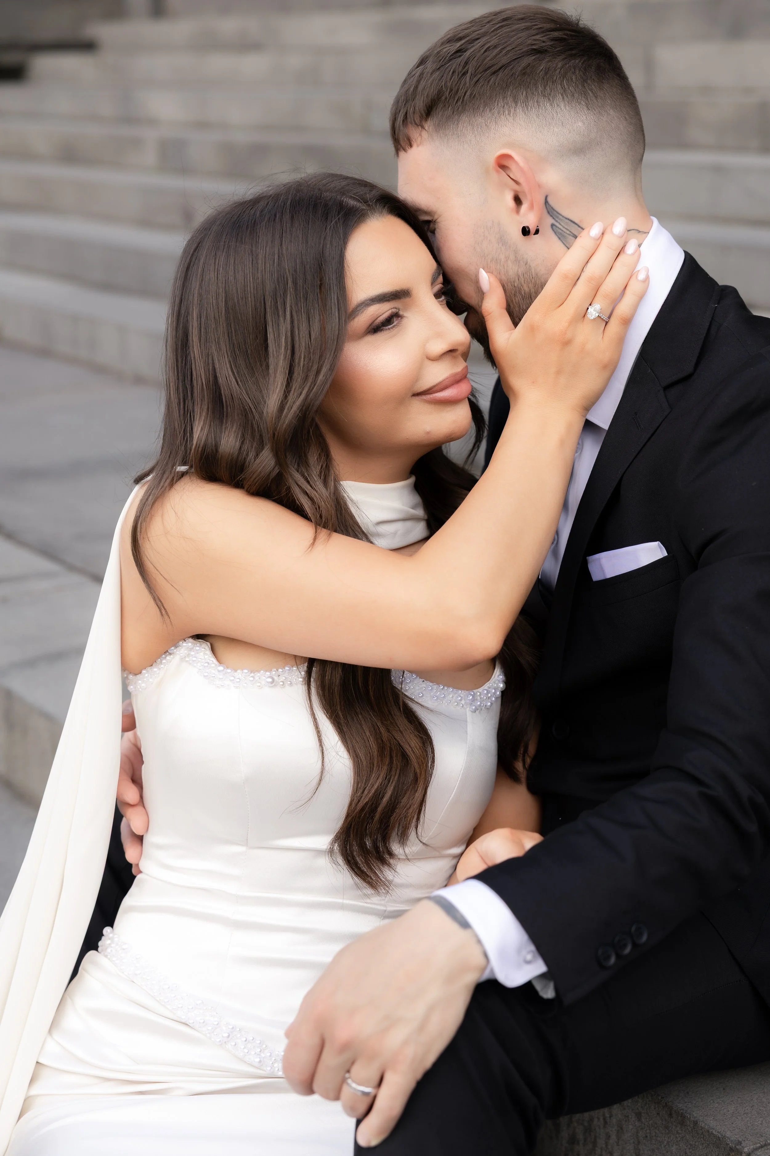 A bride and groom embracing closely, with the bride touching the groom's face and his hand on her lap, on a staircase.