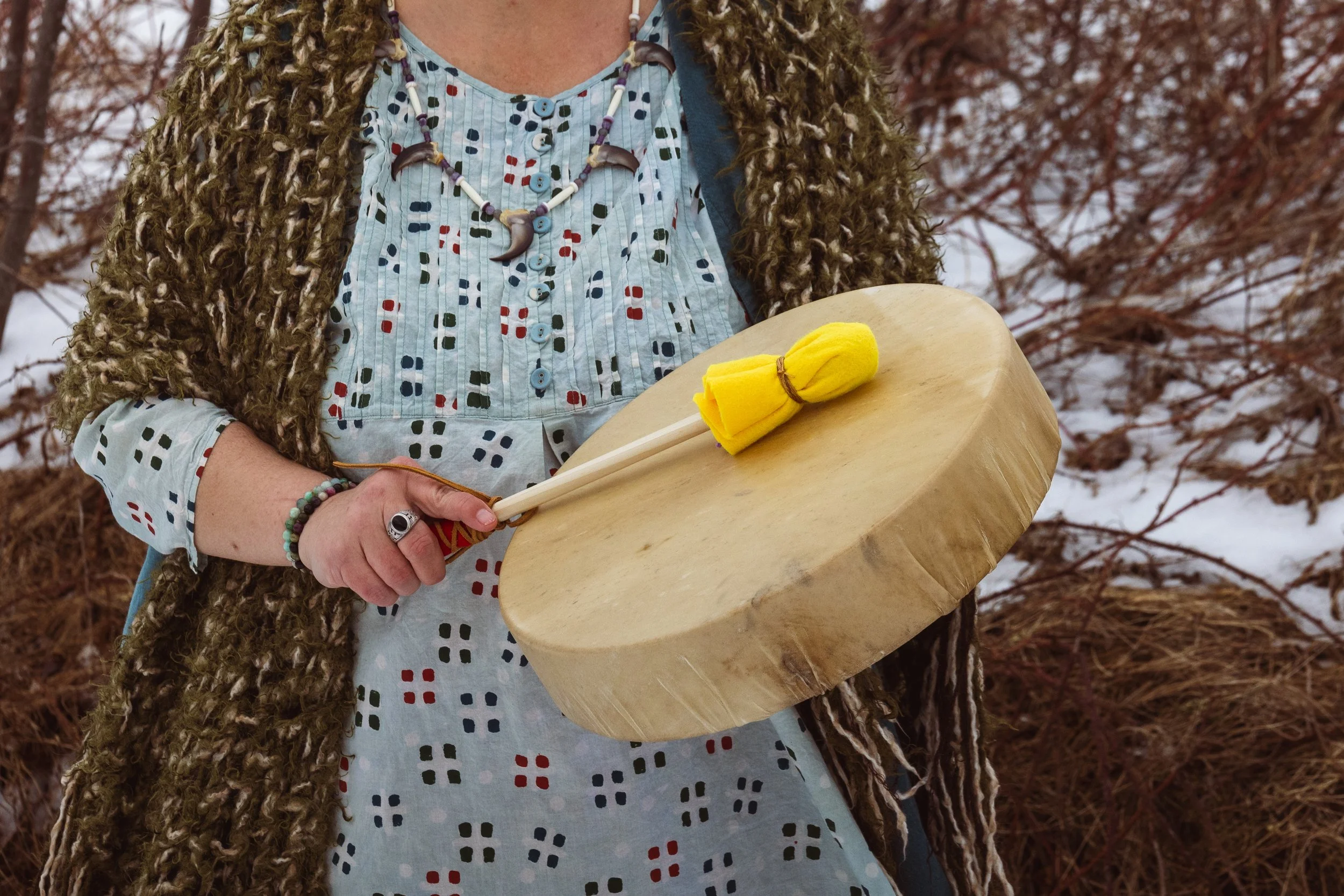 A person holding a wooden drum with a yellow felt mallet on top of it, wearing a patterned shirt, jewelry, and a furry coat, outside in a wooded area with snow on the ground.