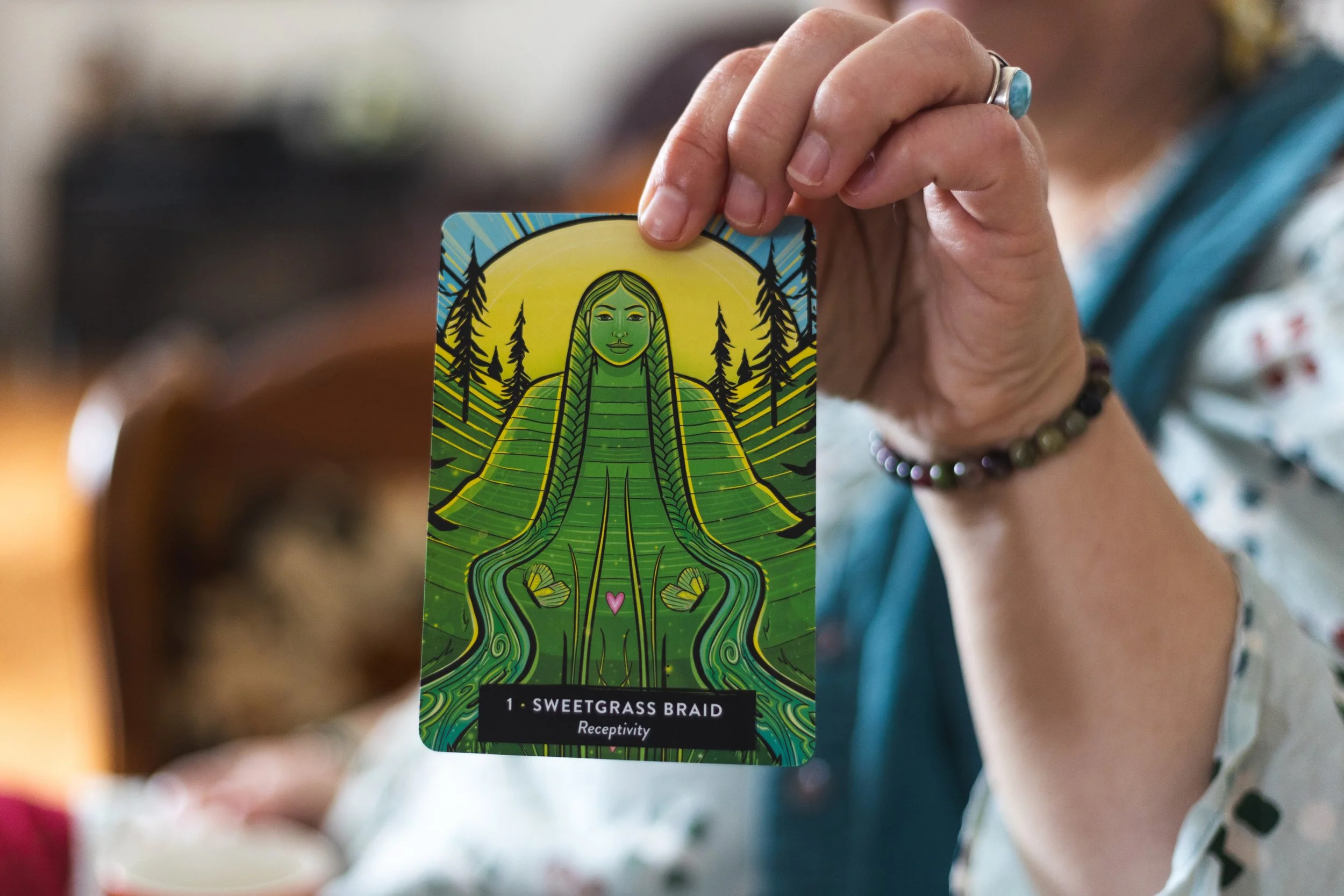Close-up of a person's hand holding a card with a colorful illustration of a woman with long hair, surrounded by trees and the moon, labeled 'Sweetgrass Braid - Receptivity'.