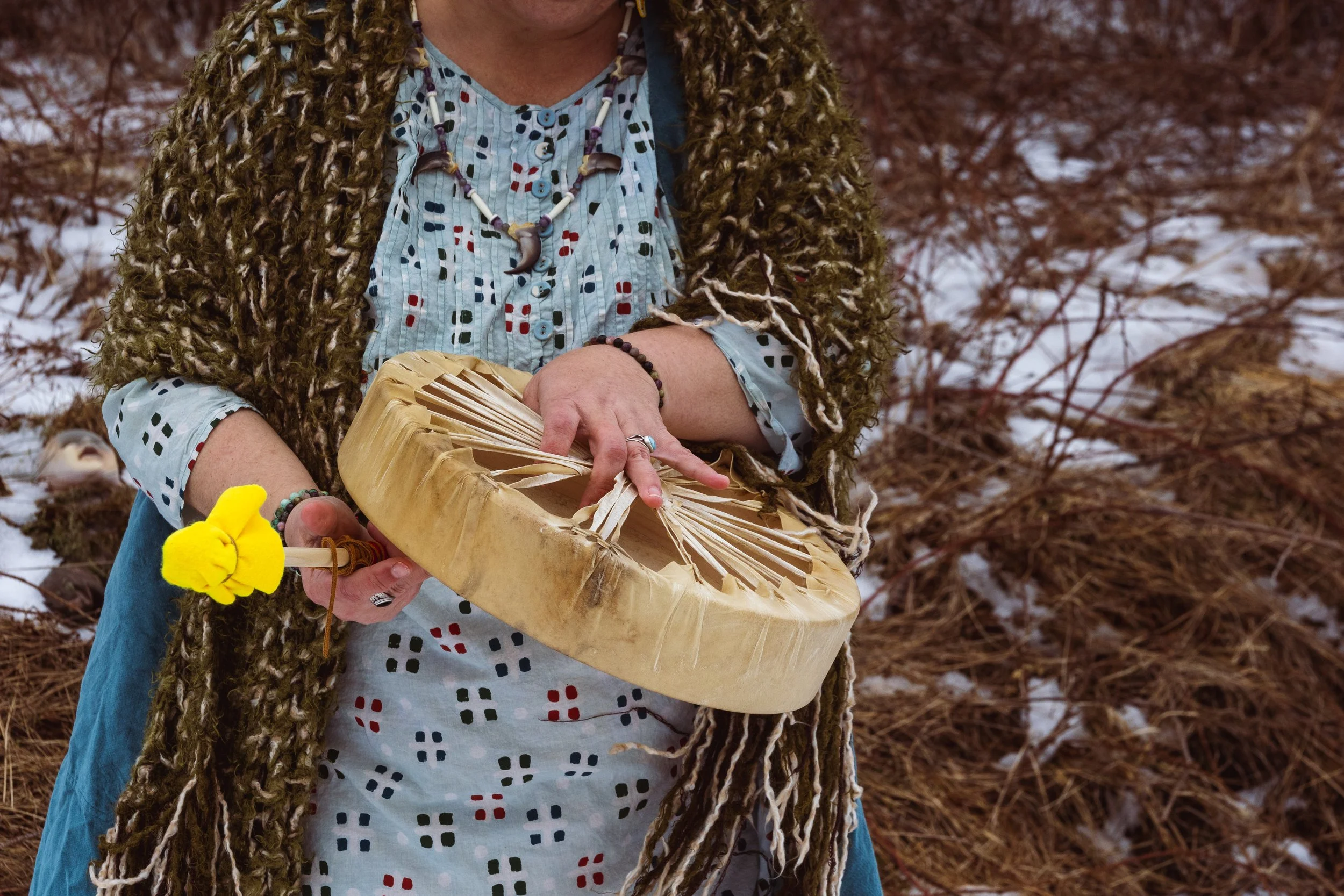 Person wearing a patterned blue shirt and a fur coat, holding a carved wooden object outdoors in a snowy, wooded area.
