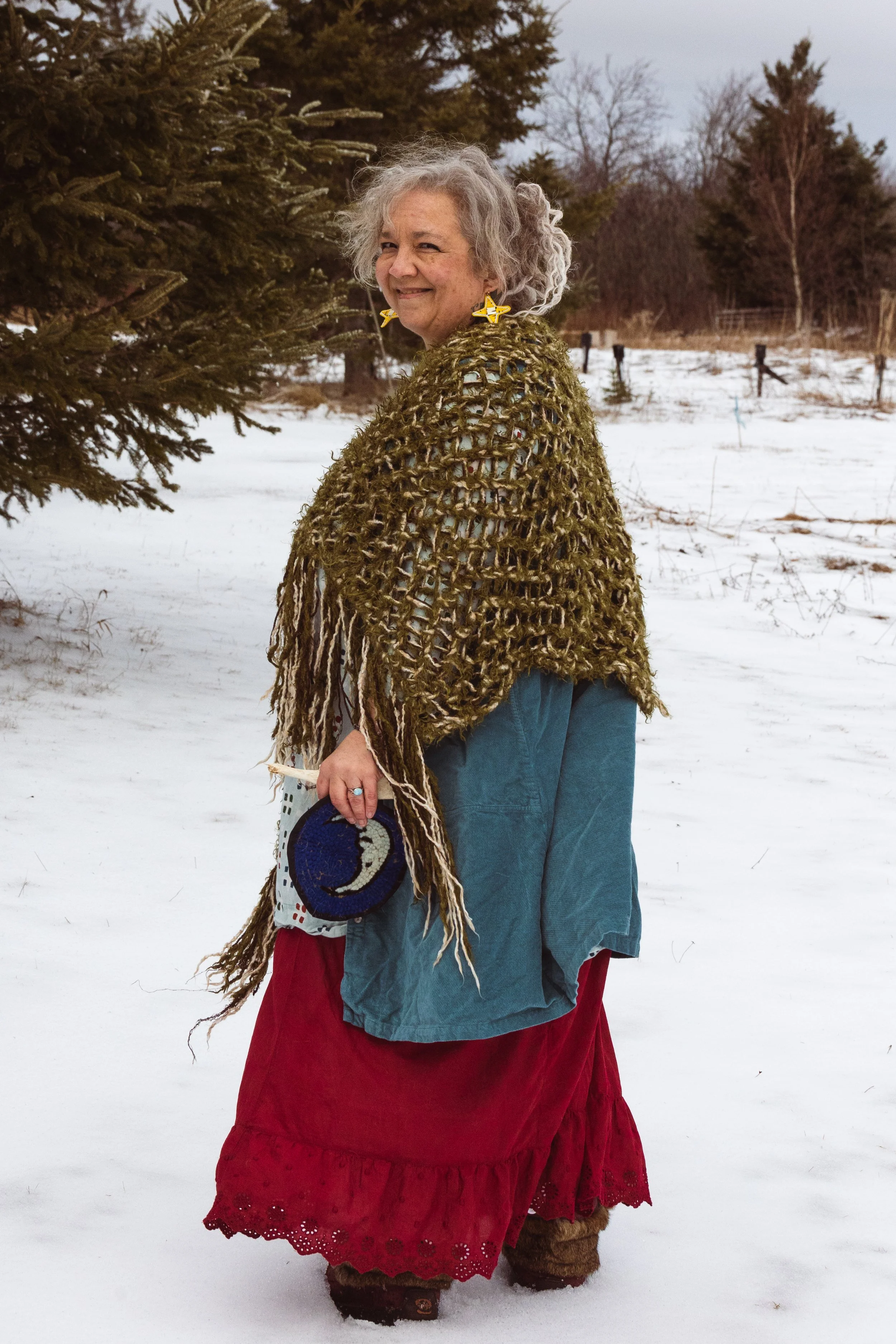 A woman standing outdoors in a snowy landscape, wearing a colorful layered outfit with a red skirt, blue coat, and a fur-lined moccasin, along with a large, crocheted shawl, and star-shaped earrings, smiling and looking over her shoulder.