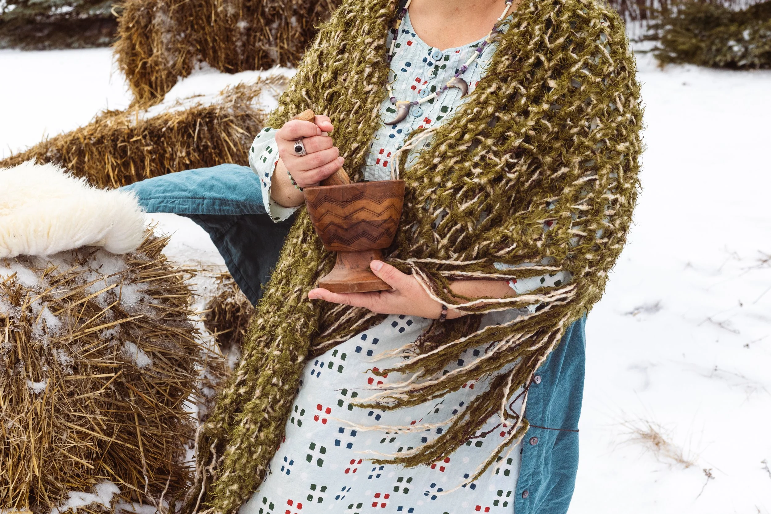 A person wearing a mossy green knitted shawl, holding a wooden chalice, standing outdoors in a snowy landscape with hay bales.
