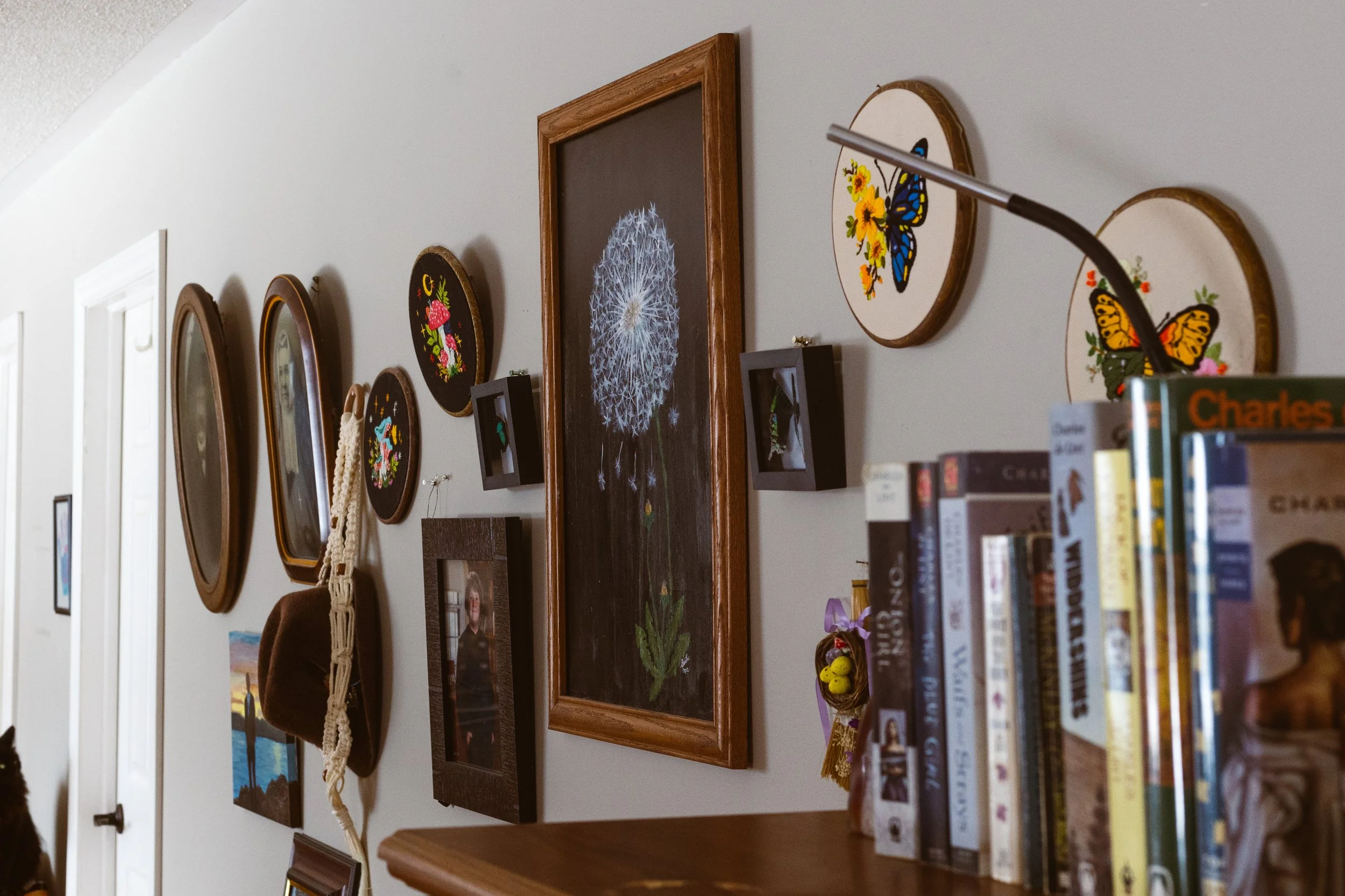 A wall decorated with various framed art, including floral and butterfly designs, with a bookshelf in the foreground.
