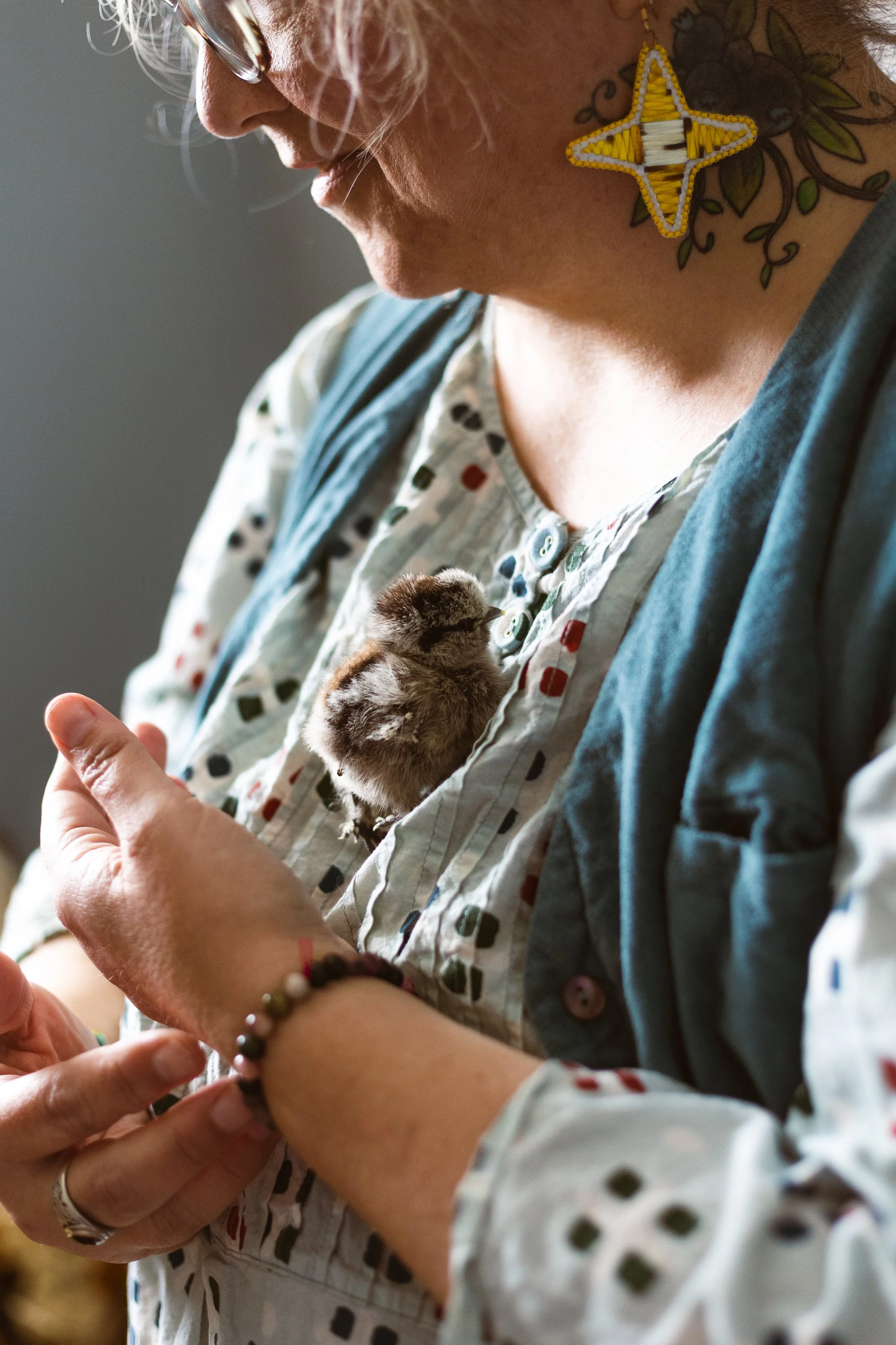 A person with glasses looking down at a small chick in their hands. They have a floral t-shirt, a beaded bracelet, and a large star-shaped beaded earring.